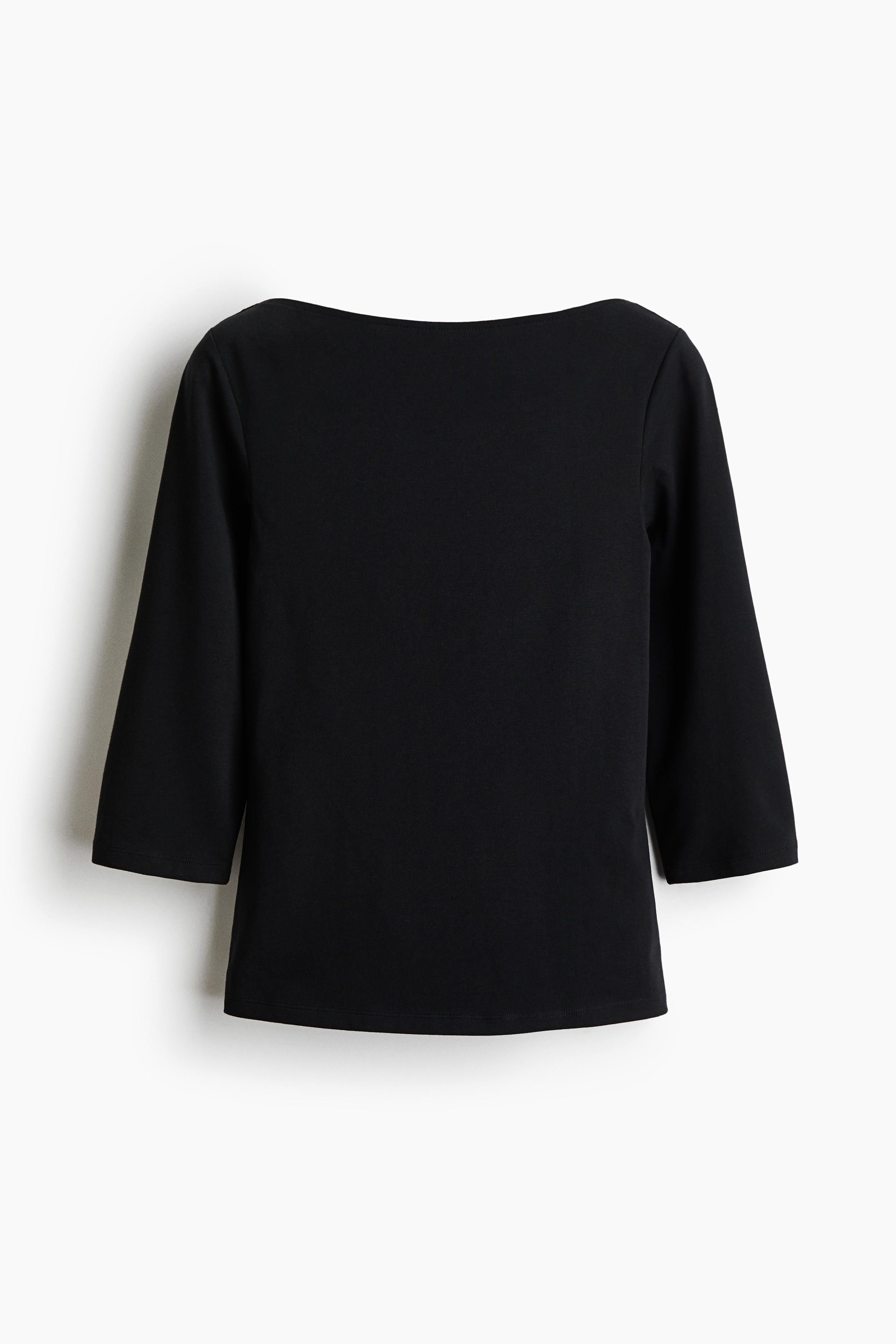 En sort, tætsiddende top i bomuldsjersey med bådudskæring og trekvartlange ærmer bæres af modellen. Modellen holder den nederste kant af toppen op med den ene hånd og afslører taljen på et par blå jeans kombineret med et sort bælte.