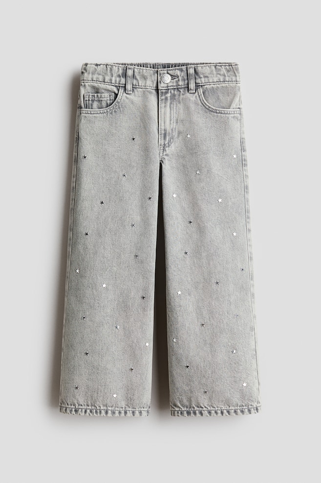 A pair of light grey wide-leg denim jeans is shown front-facing, featuring a scattered pattern of small, metallic star-shaped studs on the front legs. The jeans display a classic five-pocket design with an elasticated waistband, belt loops and a metal button fastening. - 1