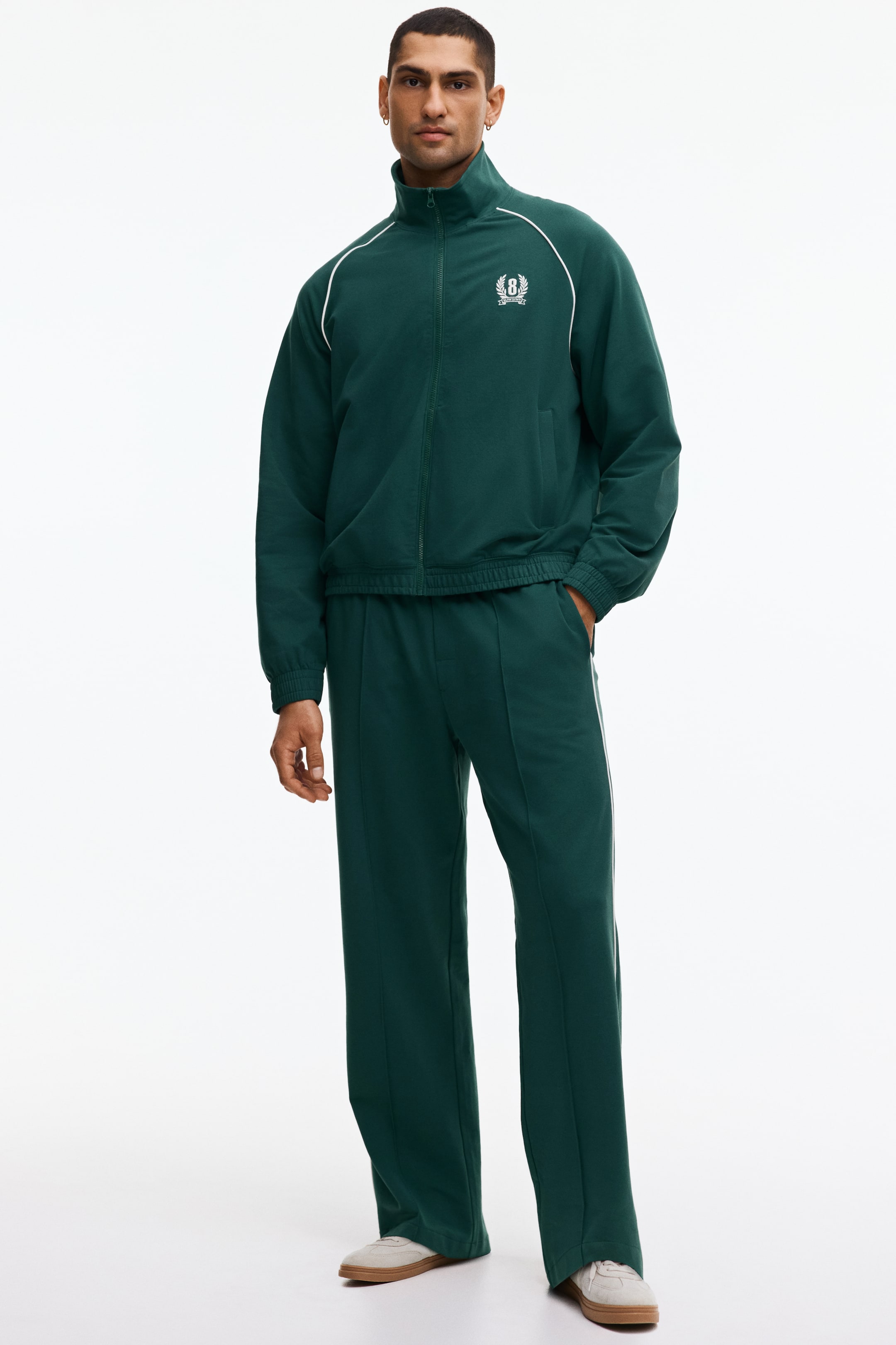 Prikaži veću sliku: Tamno zeleni trenerka komplet sadrži jaknu sa rajsferšlusom celom dužinom i uspravnom kragnom, belim paspulom duž ramena i rukava, kao i belim izvezenim amblemom na levim grudima. Jakna je završena elastičnim manžetnama i rebrastim porubom, dok odgovarajuće pantalone sa širokim nogavicama prikazuju bele bočne pruge i vertikalni prednji šav.