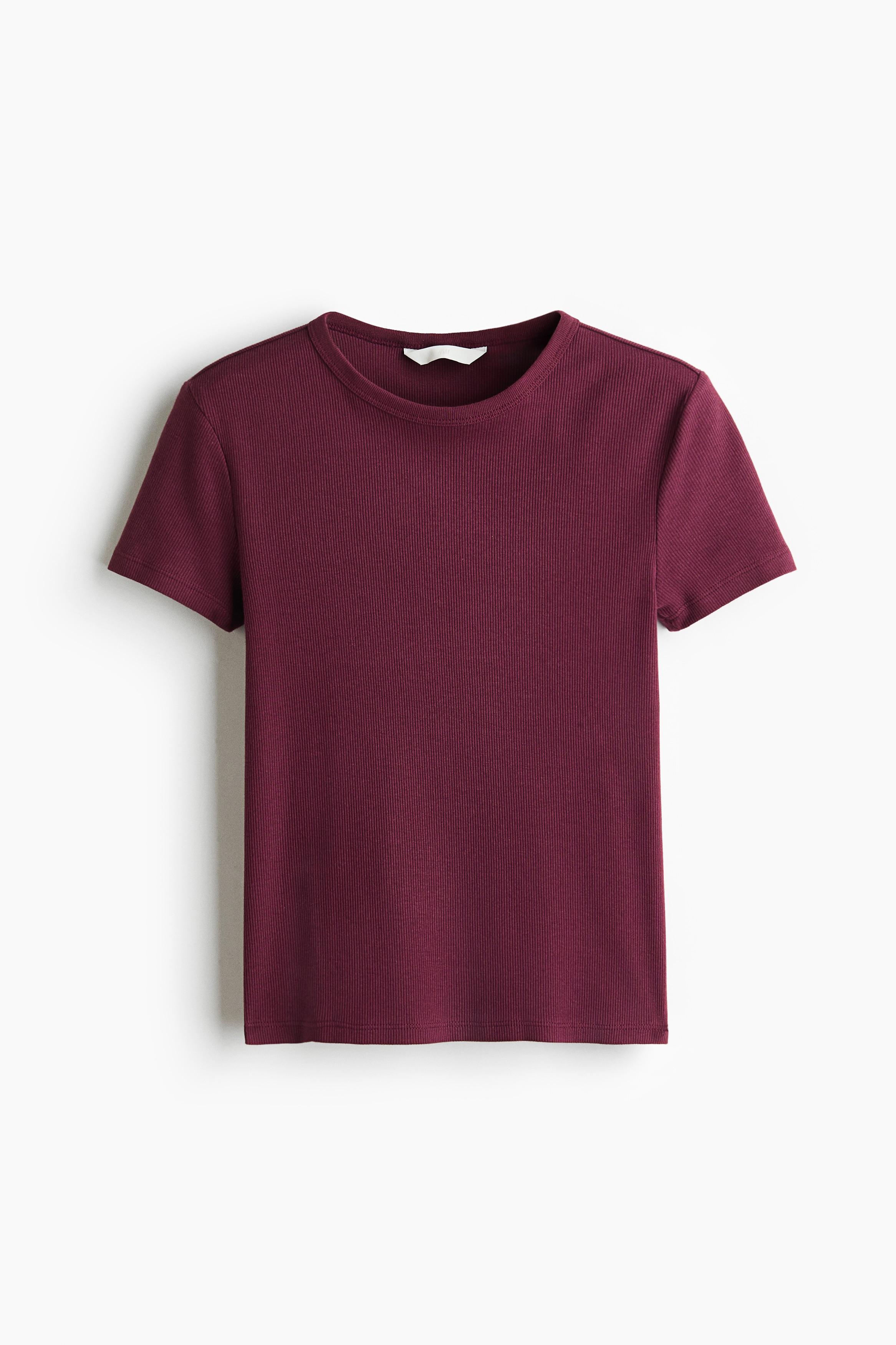 A dark red, ribbed-knit T-shirt featuring short sleeves and a narrow crew neckline is worn by the model. The model is holding the side of their waist, slightly lifting the hem of the tee to reveal the midriff above blue denim jeans.