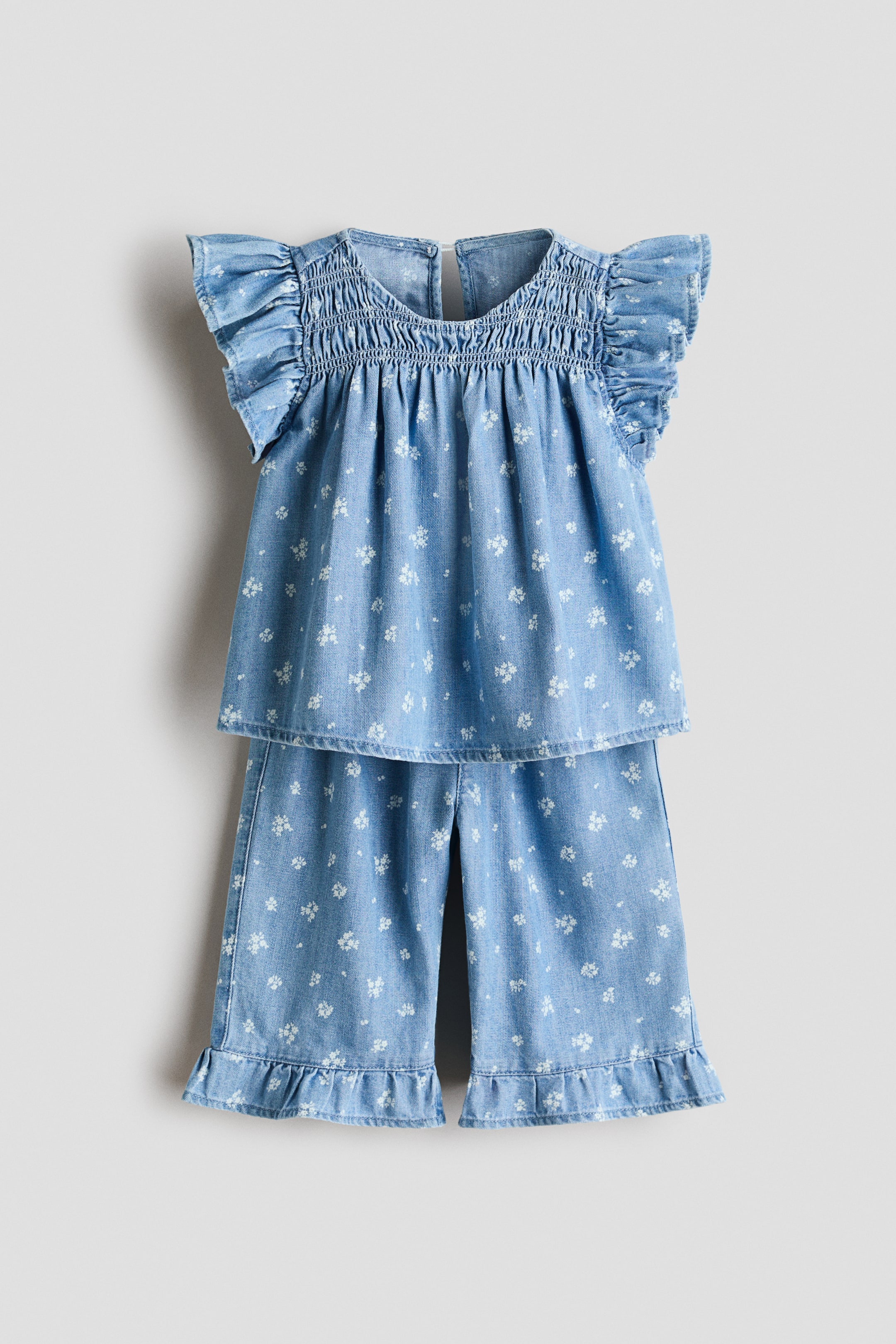 View larger image: A light blue denim two-piece set is shown facing front, featuring a white floral print throughout. The top displays a smocked bodice, a round neckline, and ruffled cap sleeves, while the matching cropped pants feature wide legs with ruffled hems.