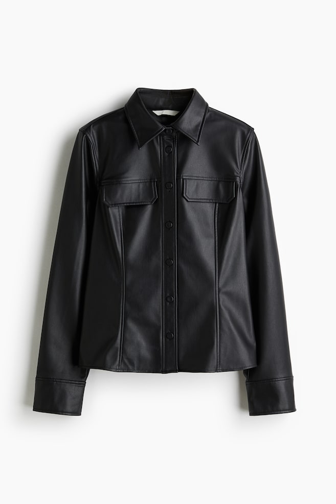 Eine schwarze Hemdjacke aus Kunstleder wird von vorne gezeigt, mit einem spitzen Kragen, einer Knopfleiste mit schwarzen Knöpfen, zwei Brusttaschen mit Knopfklappen, langen Ärmeln mit geknöpften Manschetten und vertikalen Nähten an der Vorderseite. - 1