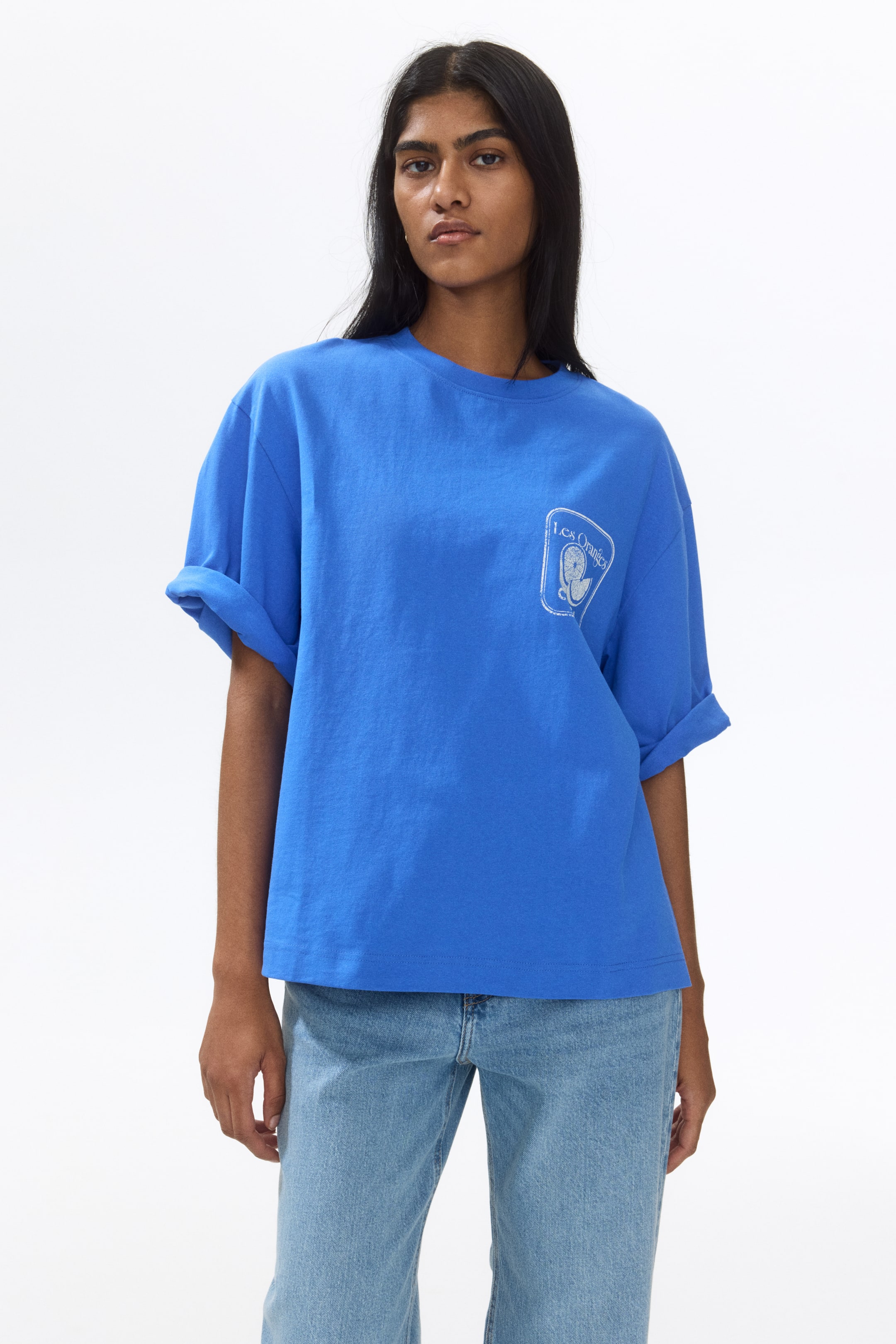View larger image: The blue t-shirt showcases a crew neck and rolled short sleeves, designed with an oversized fit. It features a white graphic on the left chest, illustrating the text "Les Oranges" alongside an orange slice design.
