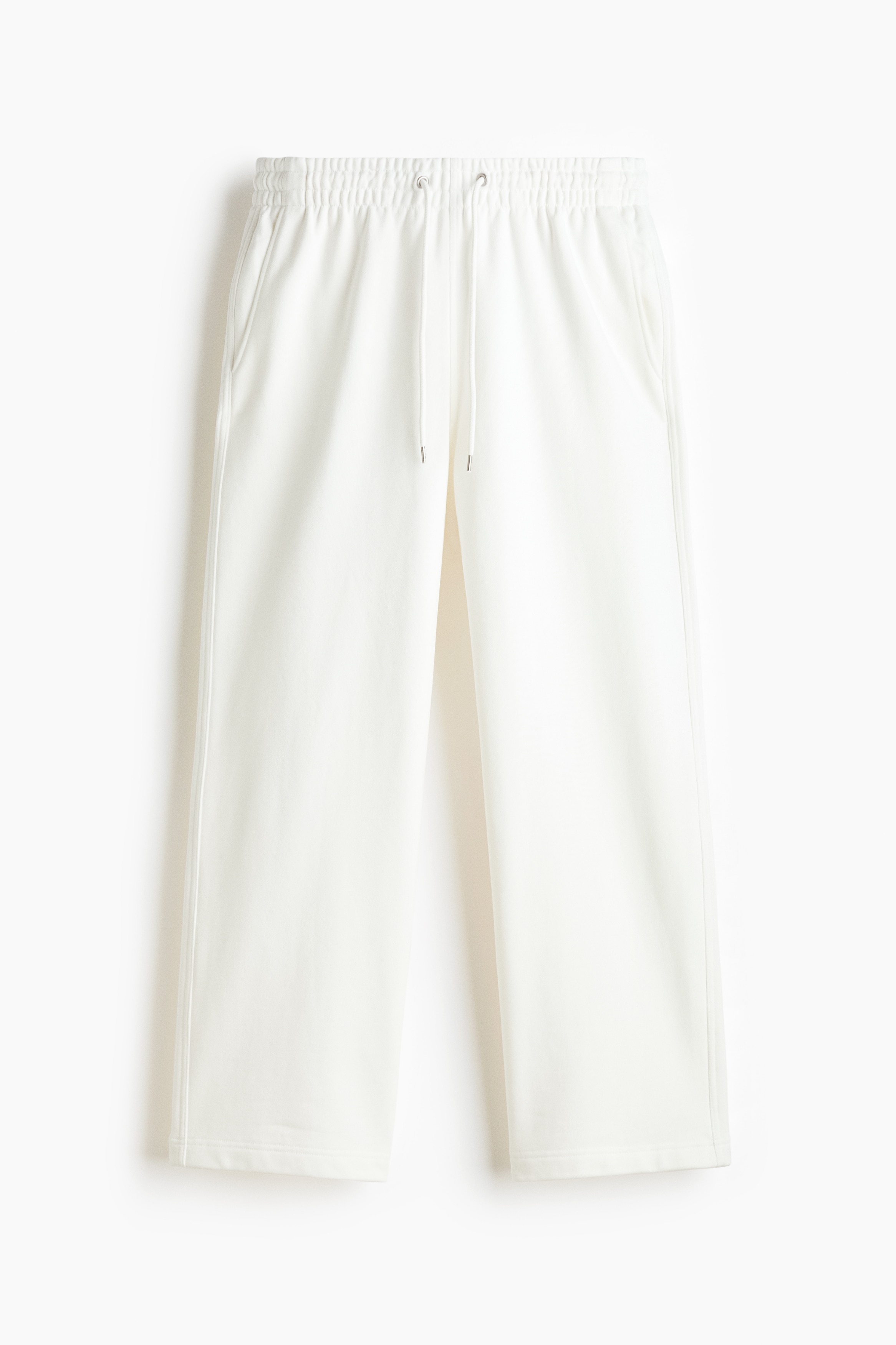 Track pants in off-white cotton sweat fabric are worn by the model, featuring a loose fit and side pockets. The model is standing with one hand resting in a side pocket, styling the trousers with a matching crewneck sweatshirt and a dark sweater tied around the waist.
