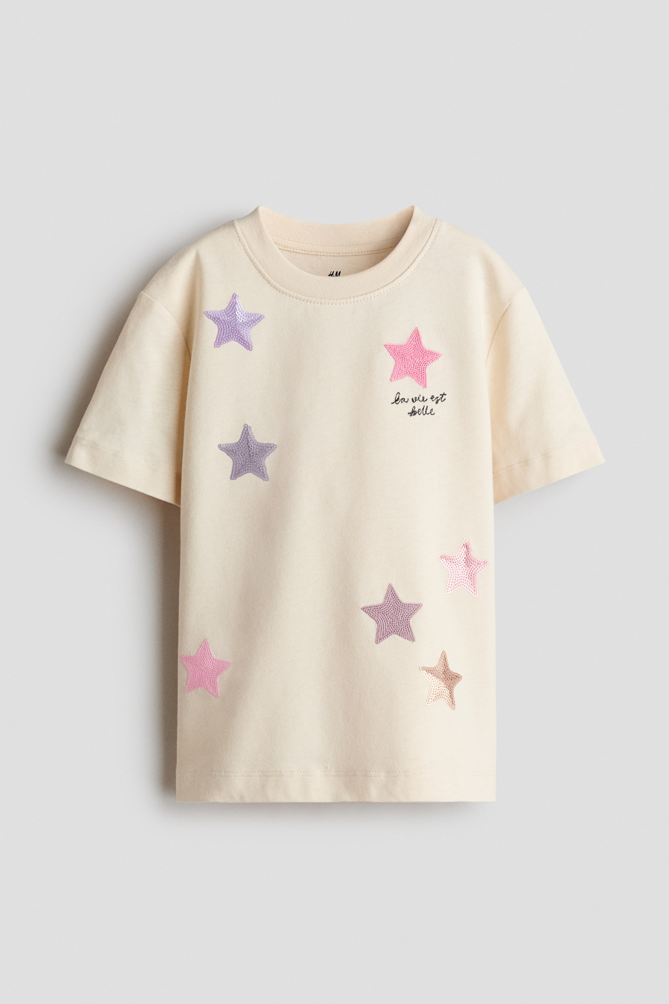 View larger image: A cream-colored t-shirt is shown from the front, featuring a ribbed crew neck and short sleeves. The front of the shirt displays scattered pastel-colored star appliqués in shades of pink, purple, and gold, along with black cursive text reading “la vie est belle” on the chest.