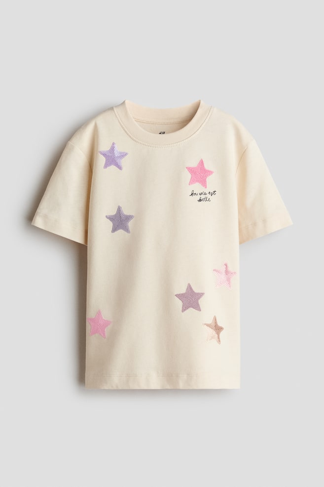 Una camiseta color crema estÃ¡ de frente, con cuello redondo de ribete acanalado y manga corta. El frente de la prenda muestra aplicaciones de estrellas en tonos pastel dispersas en rosa, morado y dorado, junto con texto negro en cursiva que dice "la vie est belle" en el pecho. - 1