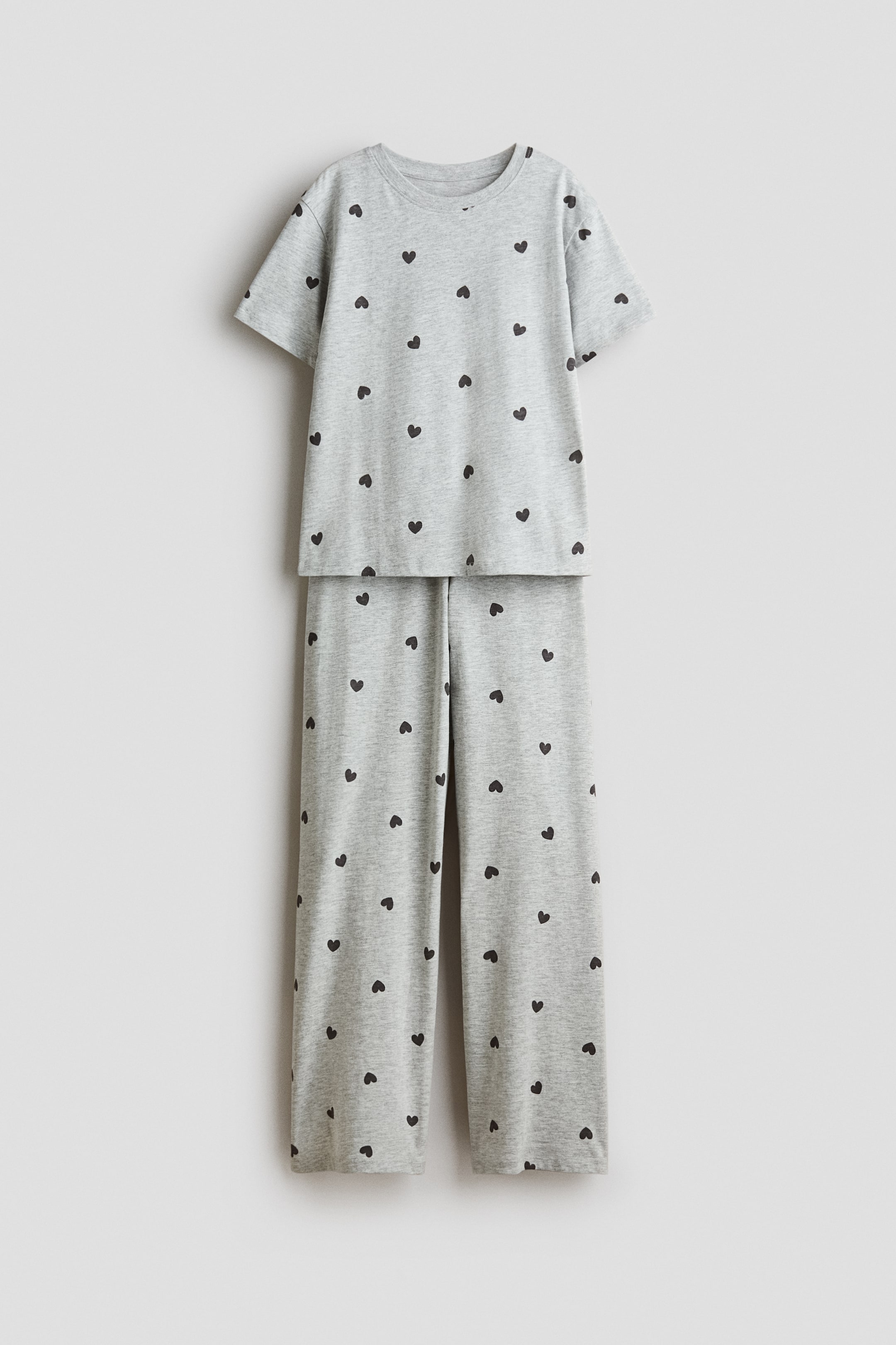 Grösseres Bild ansehen: Ein hellgraues Pyjama-Set aus Baumwolljersey zeigt eine Vorderansicht eines kurzärmeligen T-Shirts mit Rundhalsausschnitt und passender gerade geschnittener Hose, beide mit einem Allover-Muster aus kleinen schwarzen Herzen.