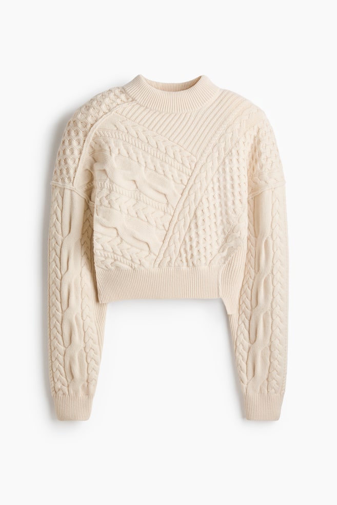 A cream-colored cropped sweater is featuring a mock neck and ribbed hem. Its body and long sleeves are displaying a complex array of cable knit patterns, including large braids, diagonal ribs, and textured diamonds, arranged in geometric panels. - 1