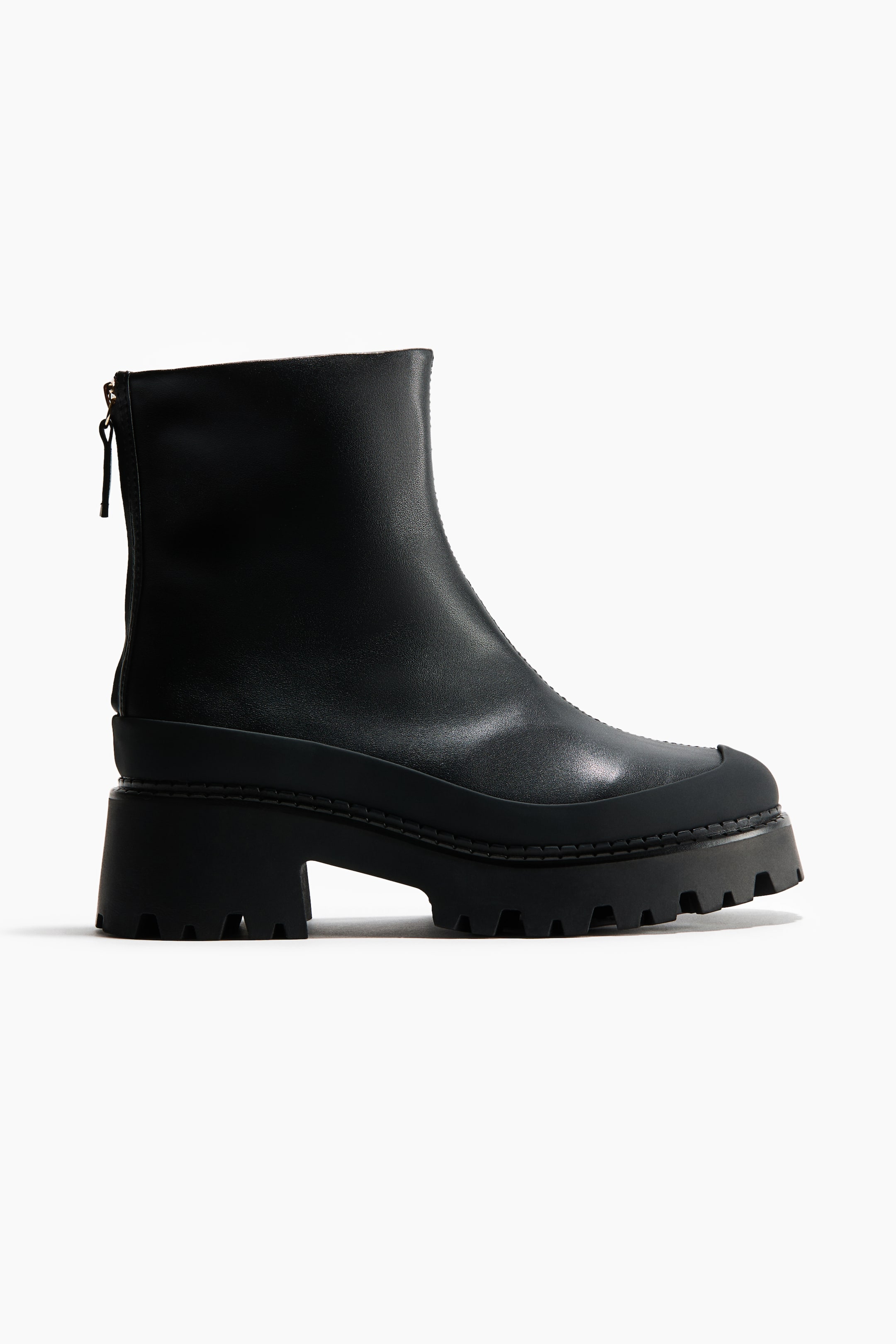 View larger image: A black leather ankle boot is shown in side profile facing right, featuring a chunky lugged rubber platform sole and a block heel. The boot is designed with a smooth leather upper, a protective rubberized mudguard wrapping around the base, and a vertical zipper closure running down the back of the heel.