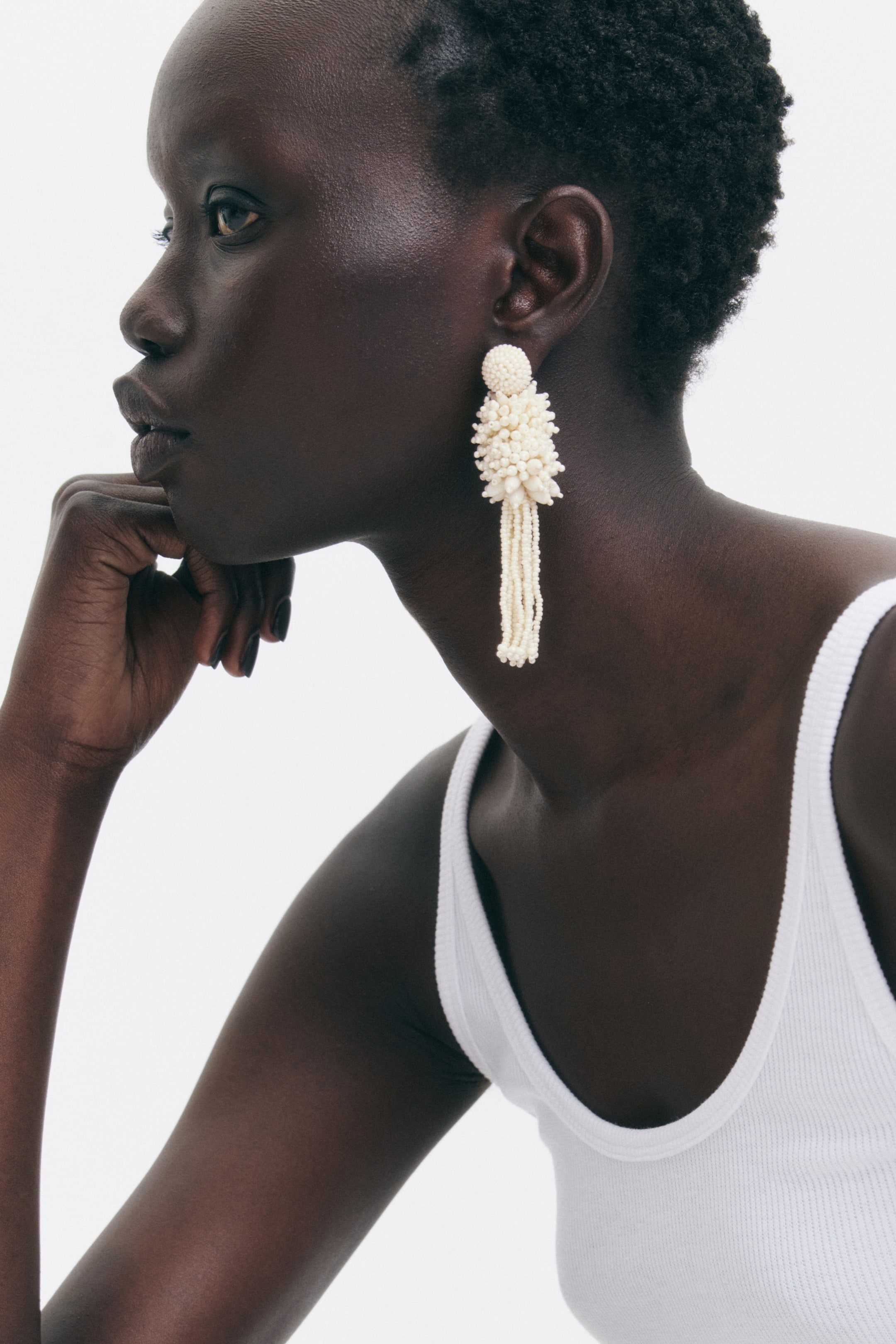 View larger image: A cream-coloured beaded earring is hanging from an ear, featuring a rounded stud, a spiky cluster of irregular beads, and multiple cascading strands forming a tassel.