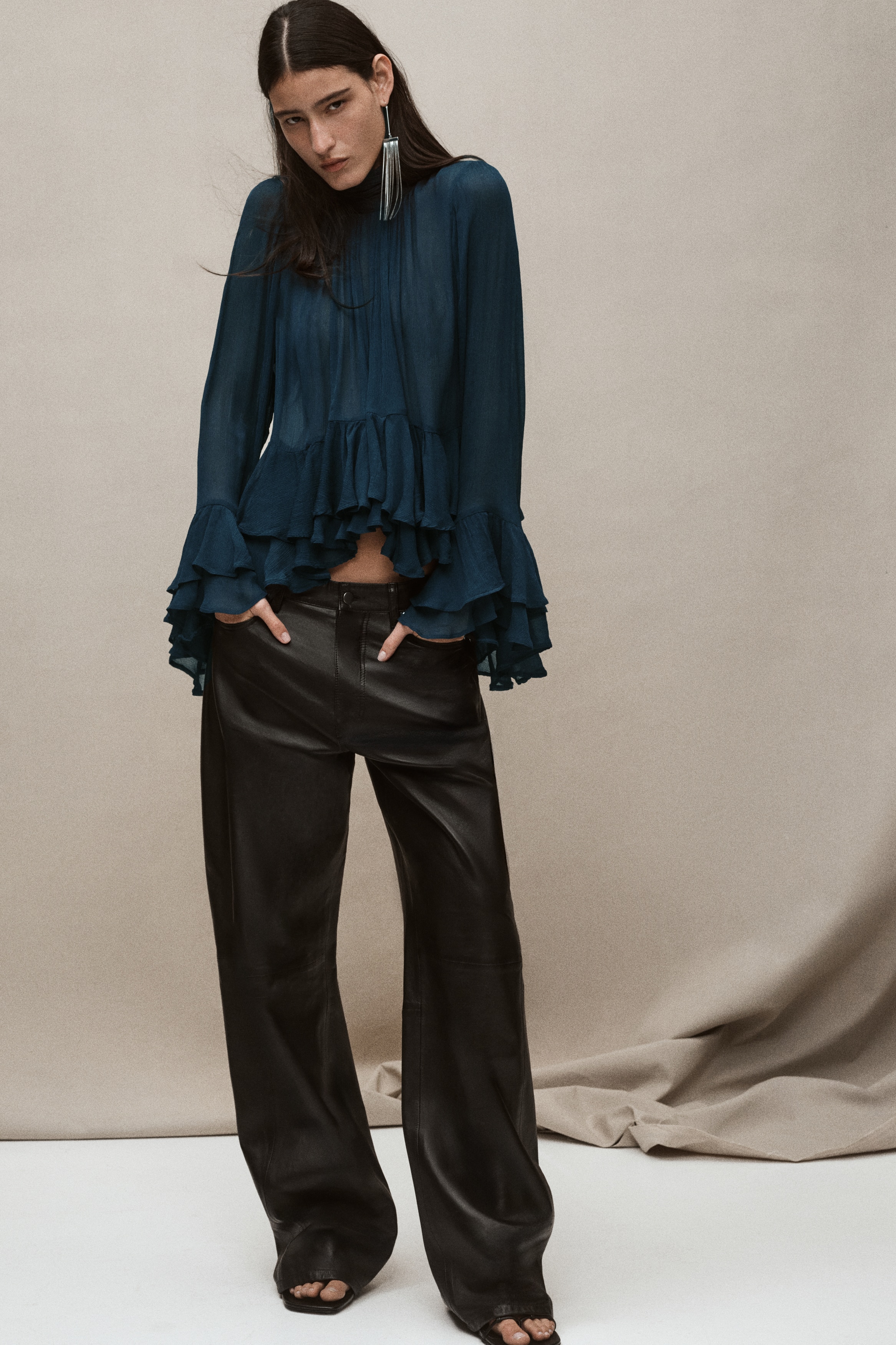 A dark teal, loose-fitting blouse is shown from the front, featuring a crinkled fabric with a pleated stand collar and a gathered bodice. The design includes long sleeves with dramatic tiered, flounced cuffs and a matching tiered peplum hem.