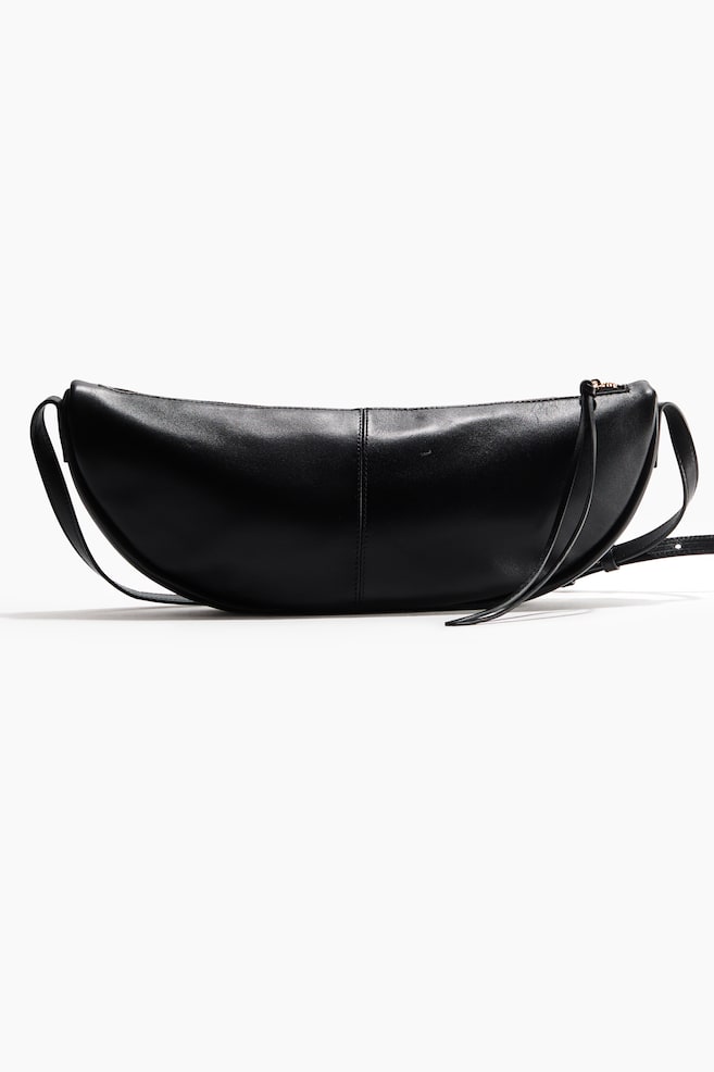 A black, crescent-shaped crossbody bag in smooth leather stands upright, facing forward. It features a central vertical seam, an adjustable shoulder strap, and a top zip fastening with a long, slim pull tab draped to one side. - 1