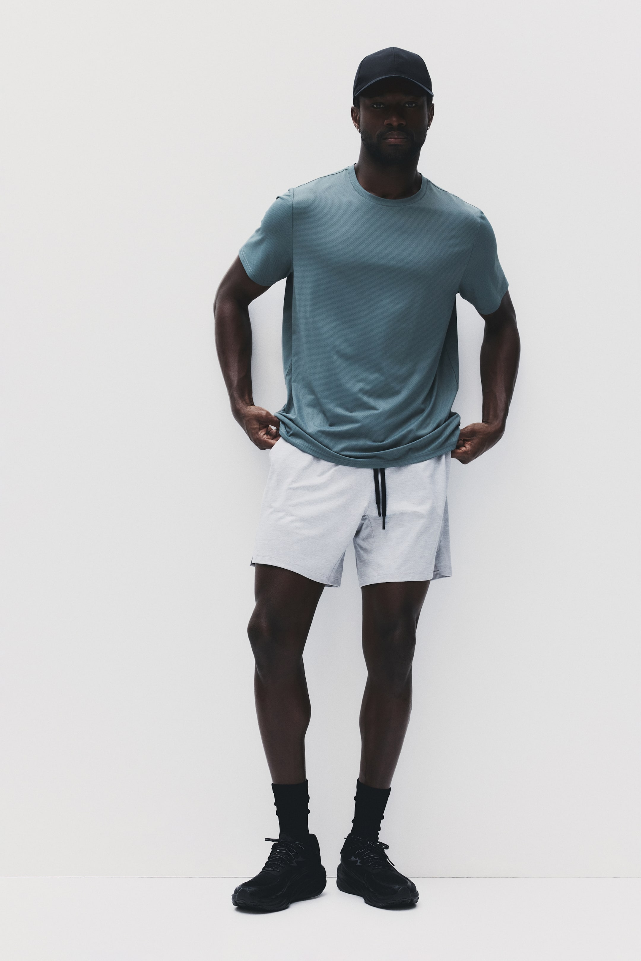View larger image: A man is donning a teal short-sleeved crew neck t-shirt with a subtle textured pattern, light grey athletic shorts featuring a black drawstring, a black baseball cap, black crew socks, and black athletic shoes with thick soles.