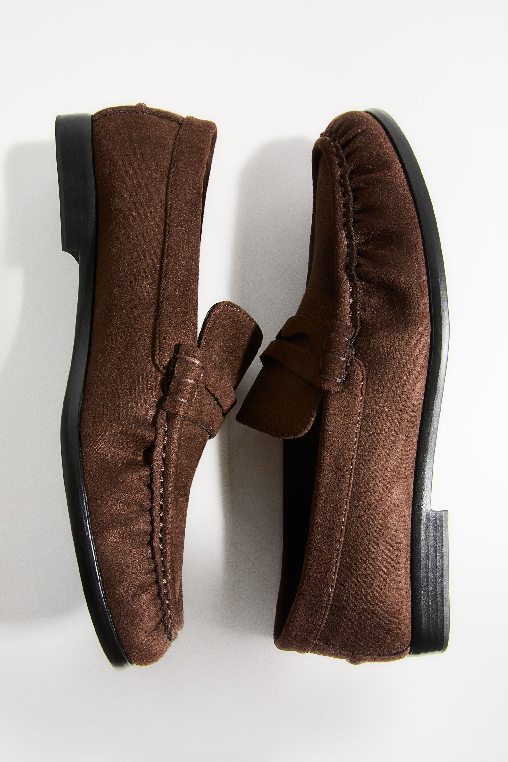 View larger image: Two brown suede penny loafers feature moccasin stitching around the toe and a classic strap across the vamp. They are presented from a top-down view, resting with their inner sides facing each other and showcasing black soles and heels.
