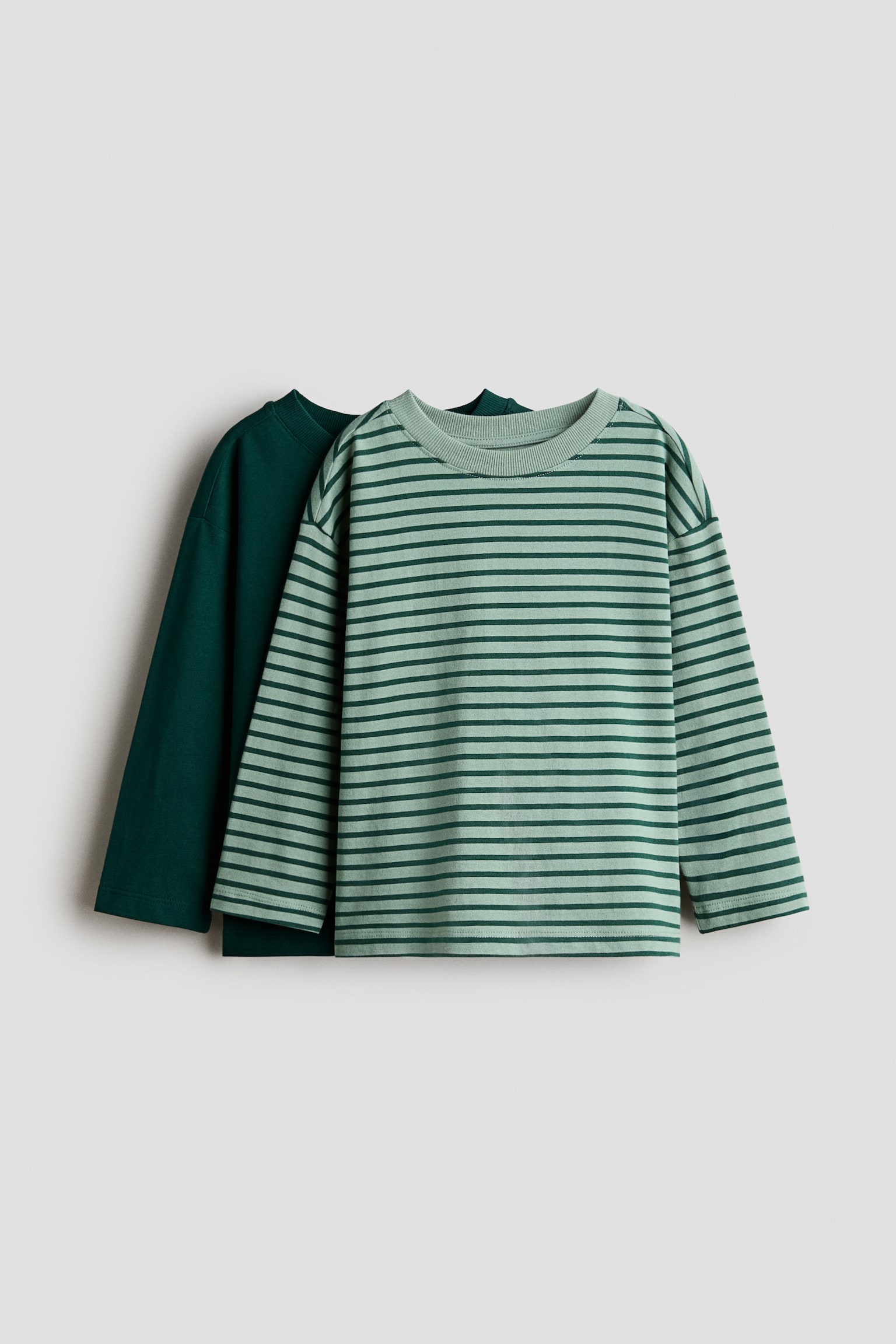 Deux t-shirts Ã  manches longues sont suspendus Ã  lâavant, lâun superposÃ© devant lâautre. Le t-shirt de devant prÃ©sente un motif rayÃ© vert clair et vert foncÃ©, avec un col rond cÃ´telÃ© et des Ã©paules tombantes, tandis que le t-shirt arriÃ¨re affiche une couleur vert foncÃ© unie.
