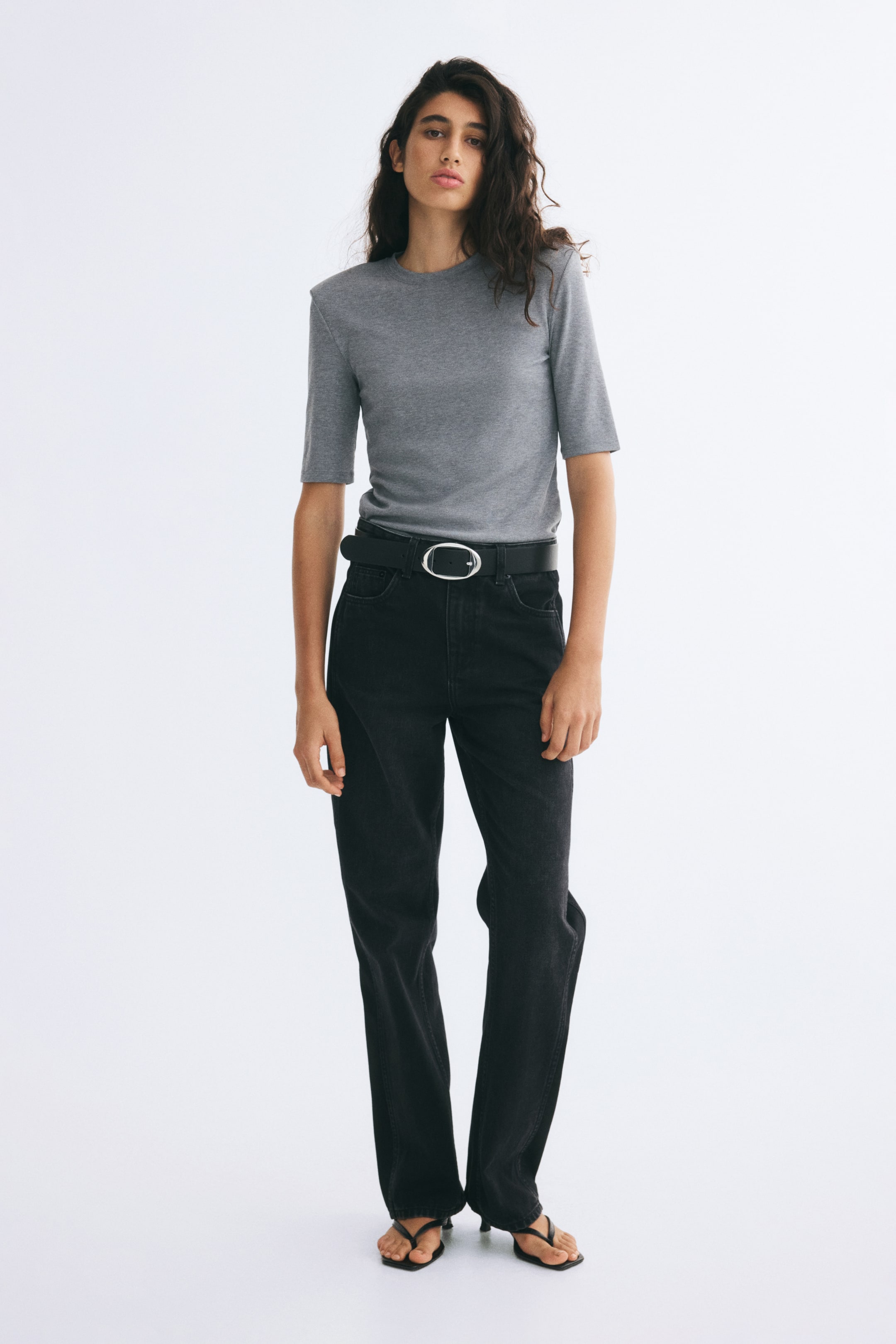 View larger image: A gray fitted tee in ribbed jersey, featuring a round neckline and structured shoulder pads, is worn by the model. The tee is styled tucked into high-waisted black jeans with a black belt.