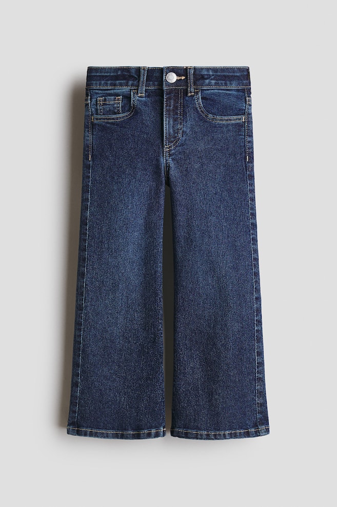 The FRONT of dark blue denim jeans is being displayed, featuring a button closure, a zip fly, and front pockets, including a small coin pocket. The jeans are presenting a slim fit through the hip and thigh, expanding into wide legs. - 1