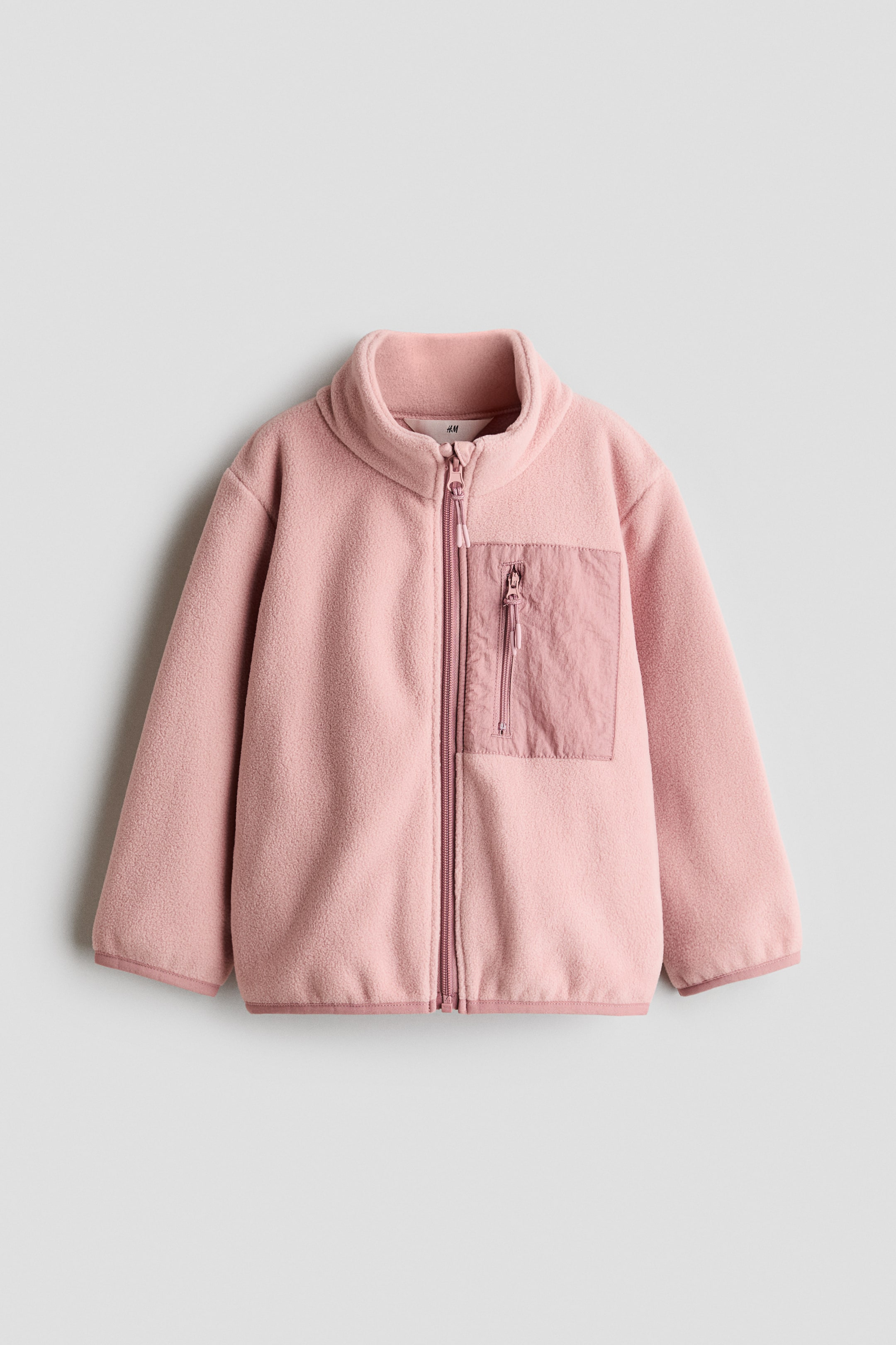 View larger image: A pink fleece jacket features a full-length pink zipper down the front and a stand-up collar. A rectangular patch pocket, made from a slightly different textured pink fabric and including a vertical zipper, appears on the left chest.