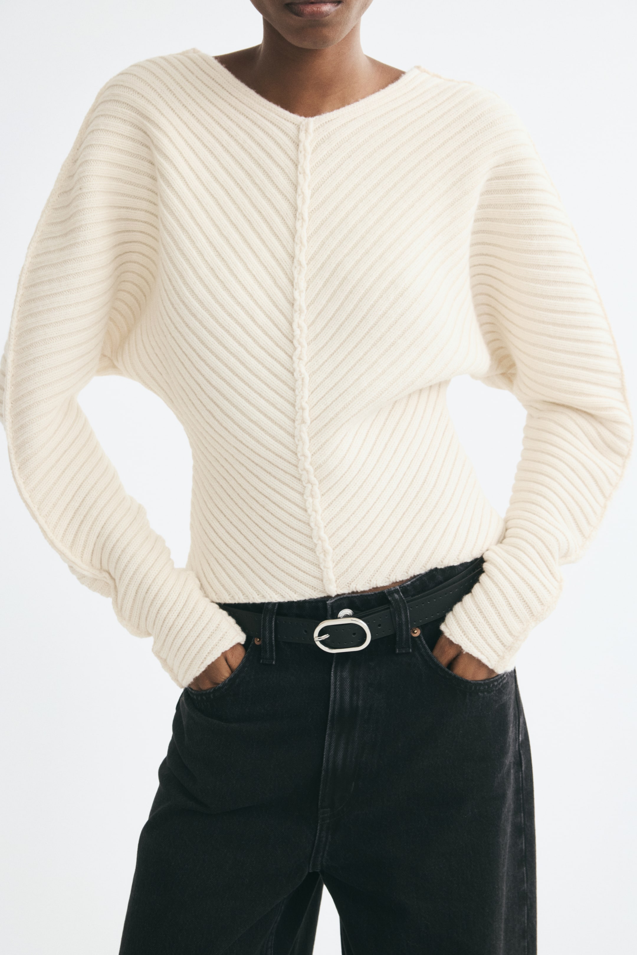 View larger image: The model is wearing an off-white rib-knit wool sweater featuring a wide V-neck, long batwing sleeves, and a central linked seam. The sweater is styled with black jeans and a black belt, and the model’s hands are resting in the pockets.