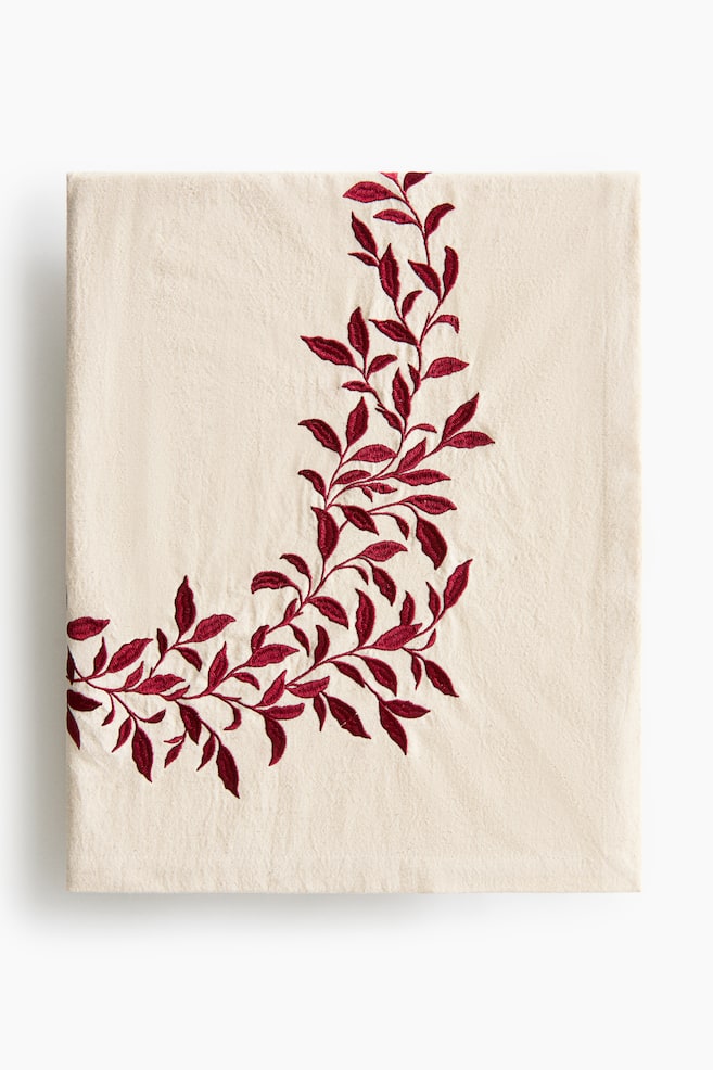 Eine gefaltete cremefarbene Stoffserviette ist frontal ausgerichtet und zeigt entlang einer Ecke ein geschwungenes, besticktes Rankenmuster mit tiefroten BlÃ¤ttern. Das Textil weist eine dezent strukturierte, gewebte OberflÃ¤che auf. - 1