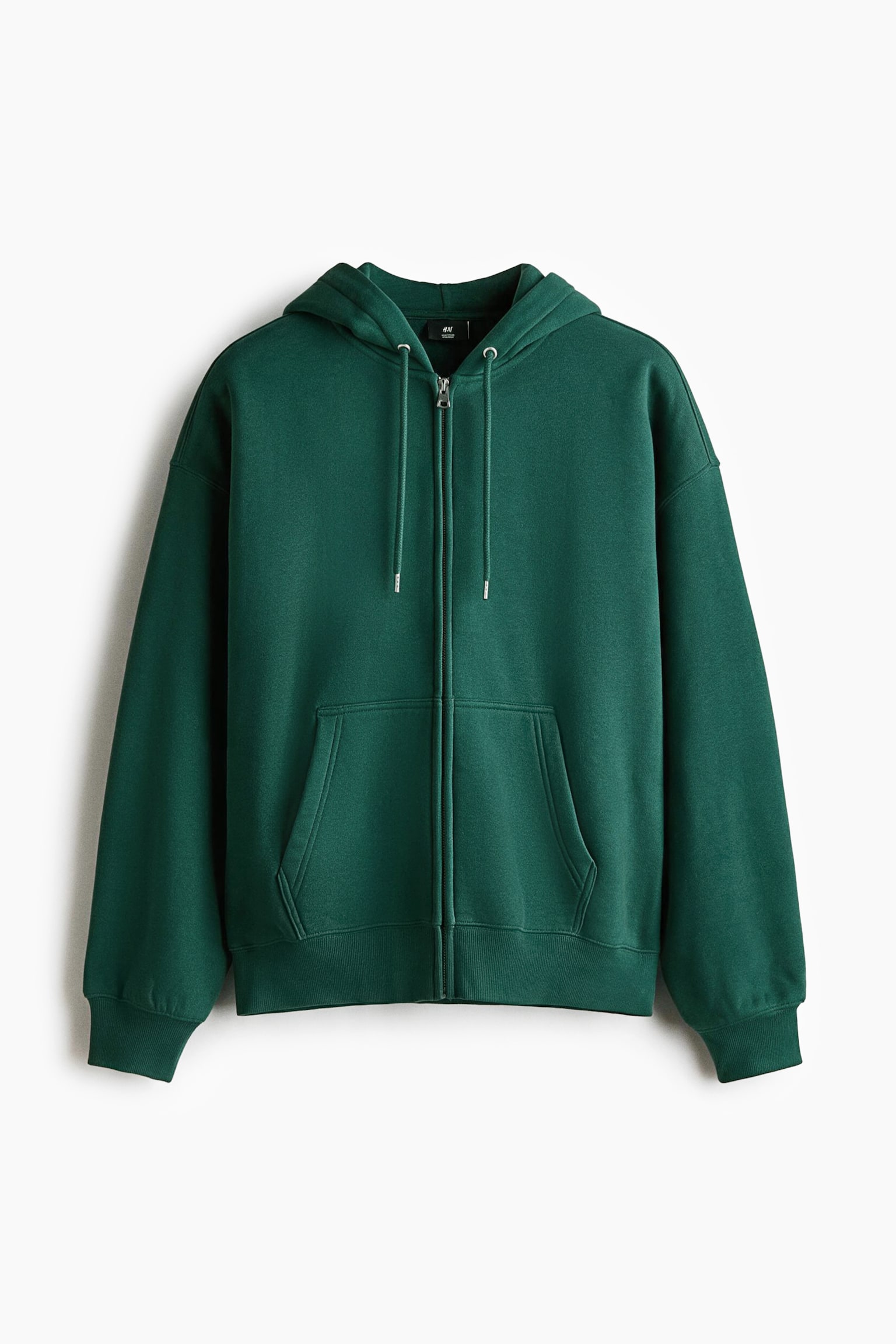 A dark green zip-up hoodie is hanging in a front-facing view, showcasing an oversized silhouette with dropped shoulders and long sleeves. It features a drawstring hood with metal-tipped cords, a full-length front zipper, split front pockets, and ribbed trim at the cuffs and hem.