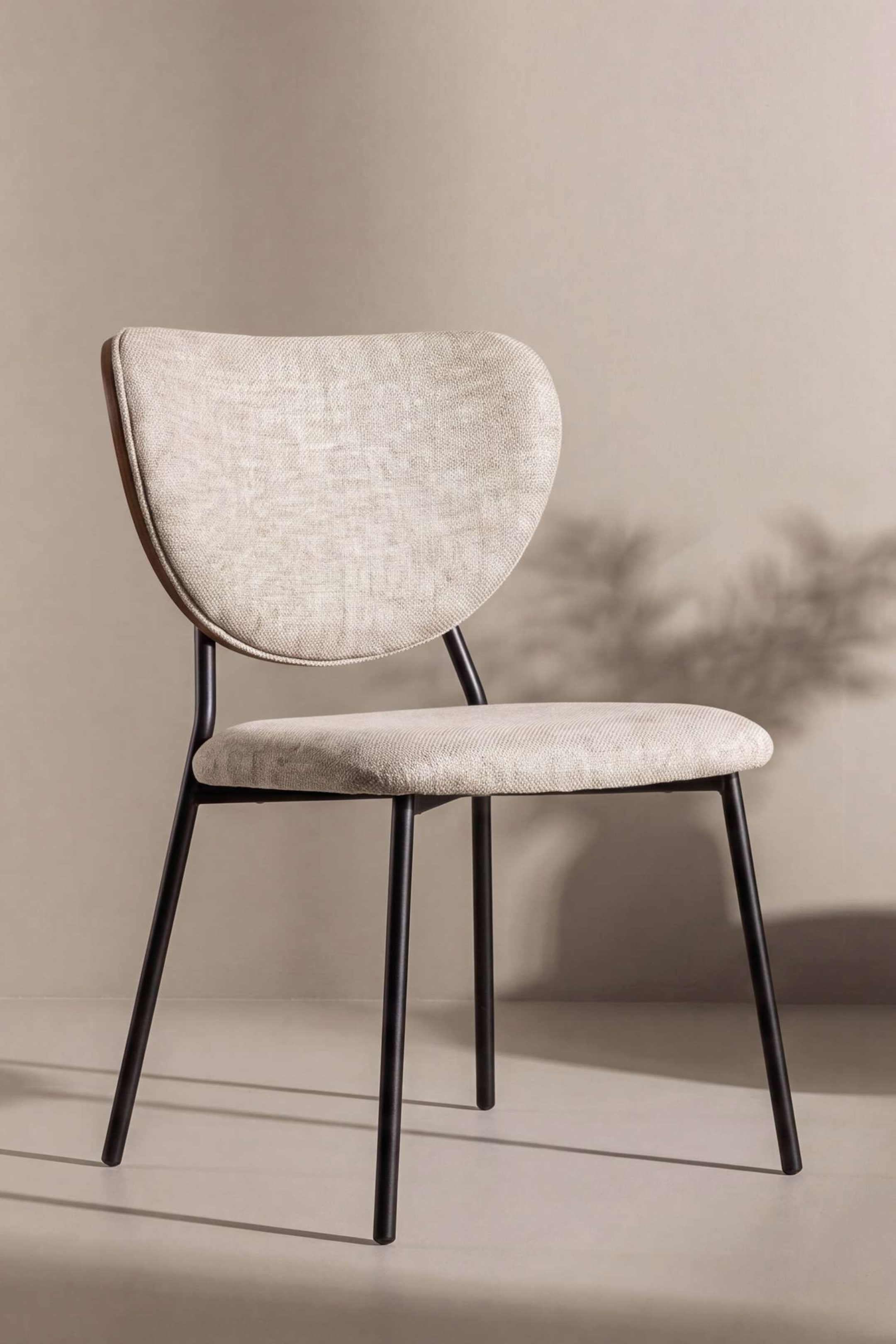 Agrandir l'image: La chaise de salle à manger Butterfly est orientée aux trois quarts vers la droite, mettant en valeur un dossier arrondi en forme de papillon avec un dossier plaqué bois foncé et une assise rembourrée, tous deux revêtus d’un tissu texturé beige clair. La chaise repose sur une fine structure en métal noir à quatre pieds fuselés.