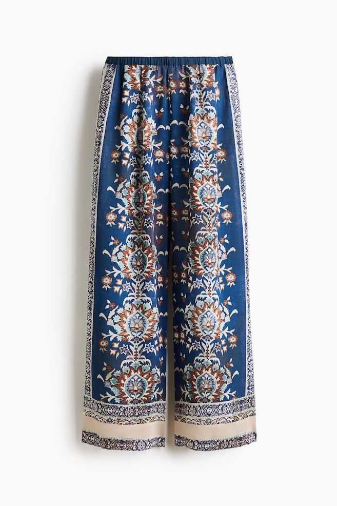 These wide-leg, loose-fit pants are shown from the front, featuring a high, elasticated waistband and an ornate floral and border pattern in blue, white, and brown. The pants are displaying wide legs with a contrasting cream-colored patterned band at the hems. - 1
