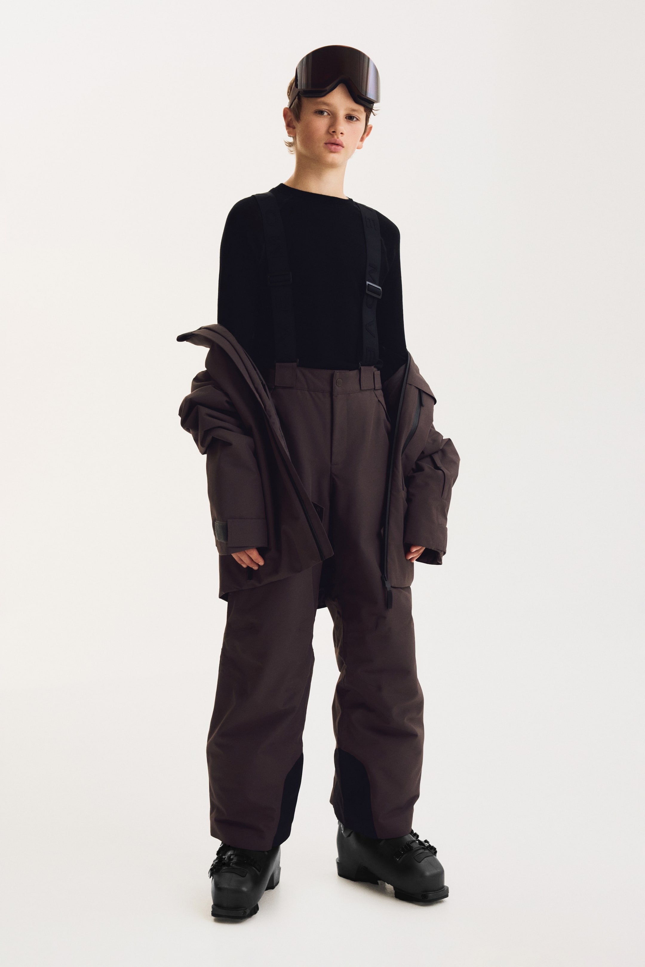 View larger image: A model is standing and facing forward, wearing a dark brown ski outfit that includes high-waisted ski pants with adjustable black suspenders and a matching hooded jacket draped off the shoulders. The ski pants feature black reinforced inner-ankle panels and are worn over black ski boots and a black long-sleeve base layer.