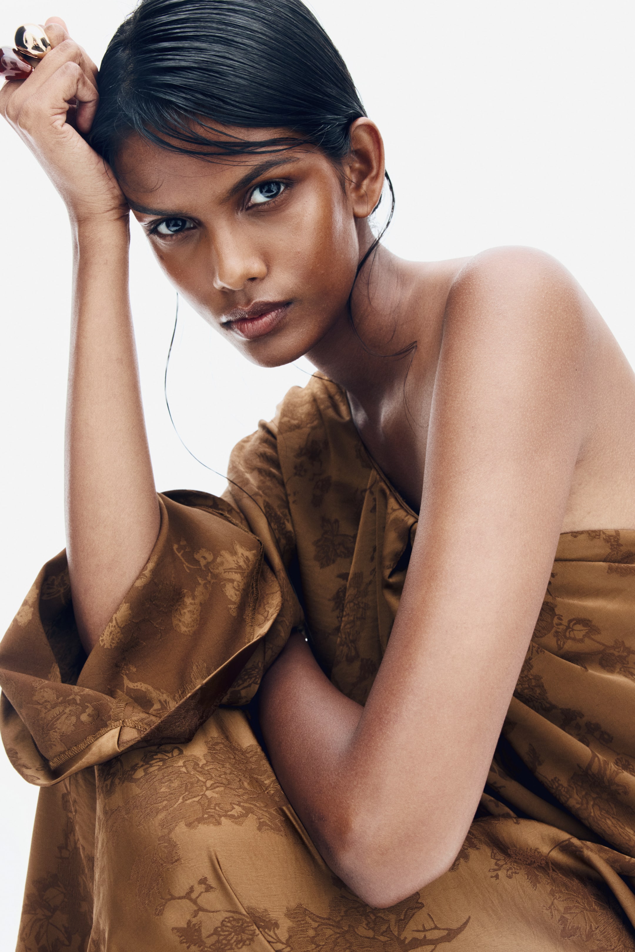 View larger image: The model is wearing a bronze-coloured, one-shoulder dress in floral jacquard weave satin, leaning forward with one arm raised to her head. The dress features an asymmetrical neckline that leaves one shoulder bare, while the draped, patterned fabric covers the other side.