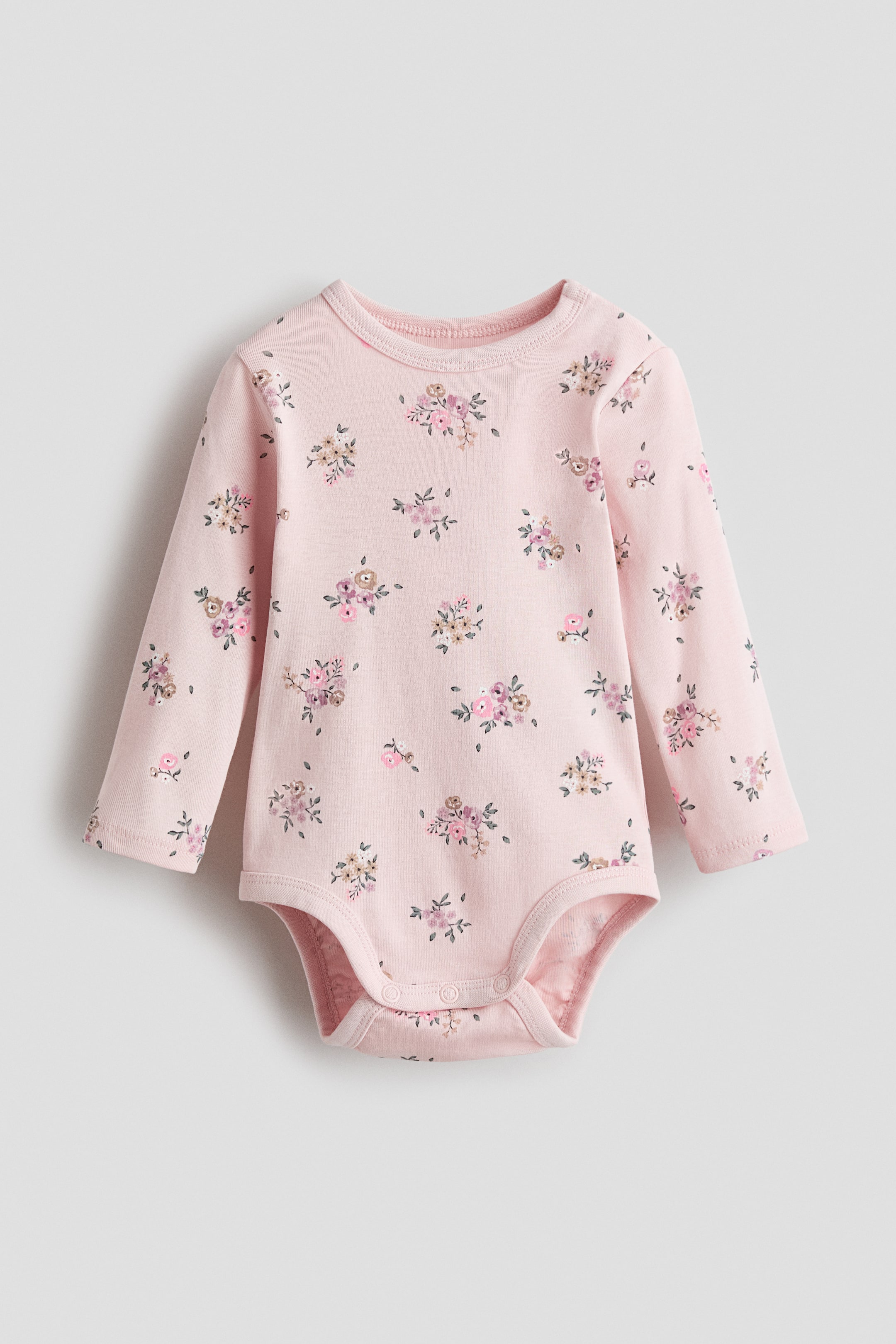 이미지 확대 보기: 앞면을 보여주는 핑크 코튼 저지 바디수트로, 핑크, 퍼플, 베이지, 그린 색상의 꽃무늬가 흩어져 있습니다. 라운드 트림 넥라인, 오른쪽 어깨의 프레스 스터드, 긴 소매, 아랫부분에 숨겨진 프레스 스터드가 디자인되어 있습니다.