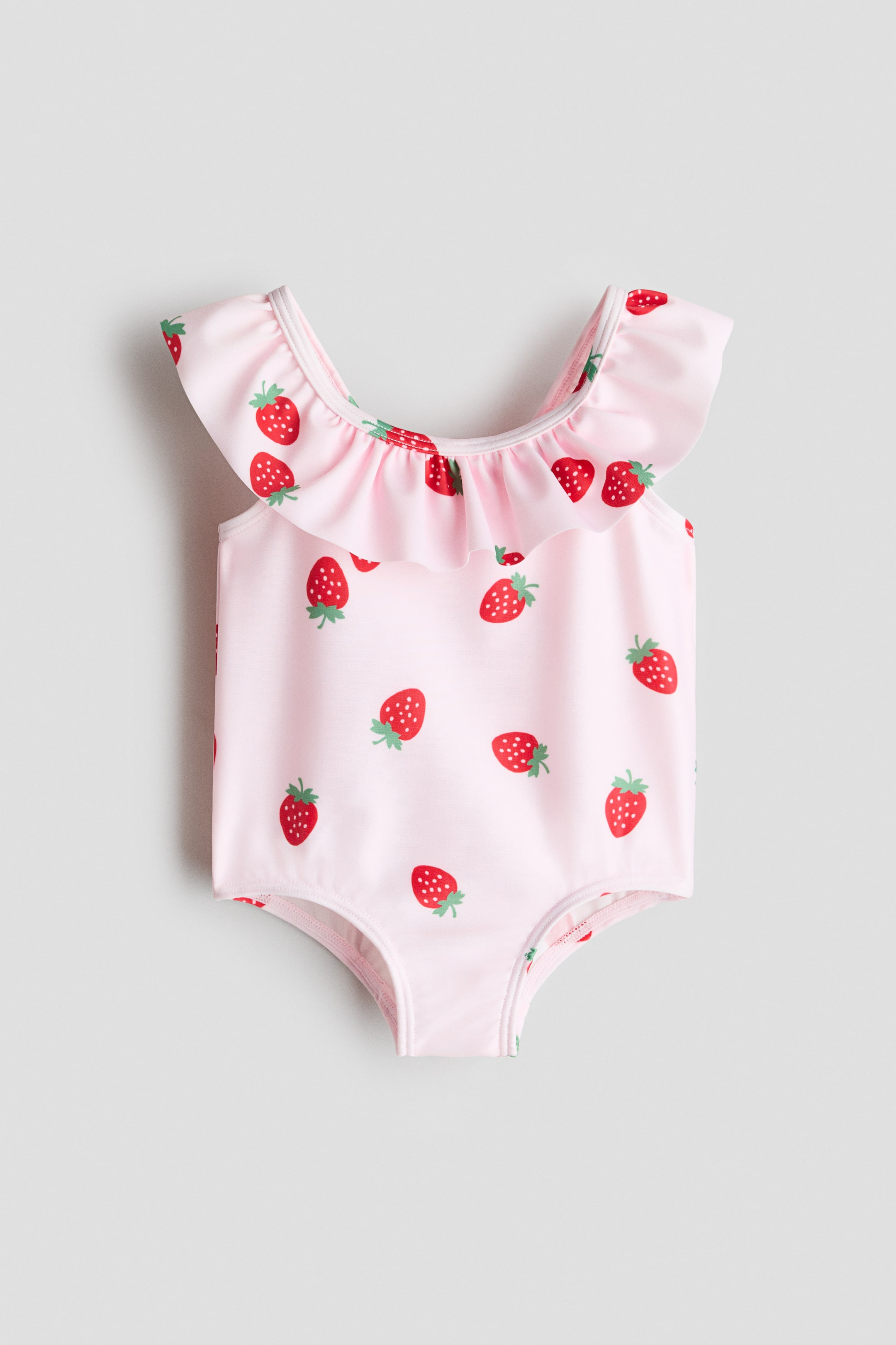 Ver imagen más grande: Un bañador de frente presenta un color rosa claro con un estampado disperso de fresas rojas. Un volante recorre el escote y se extiende sobre los hombros para formar los tirantes; un refuerzo forrado es visible en la parte inferior.