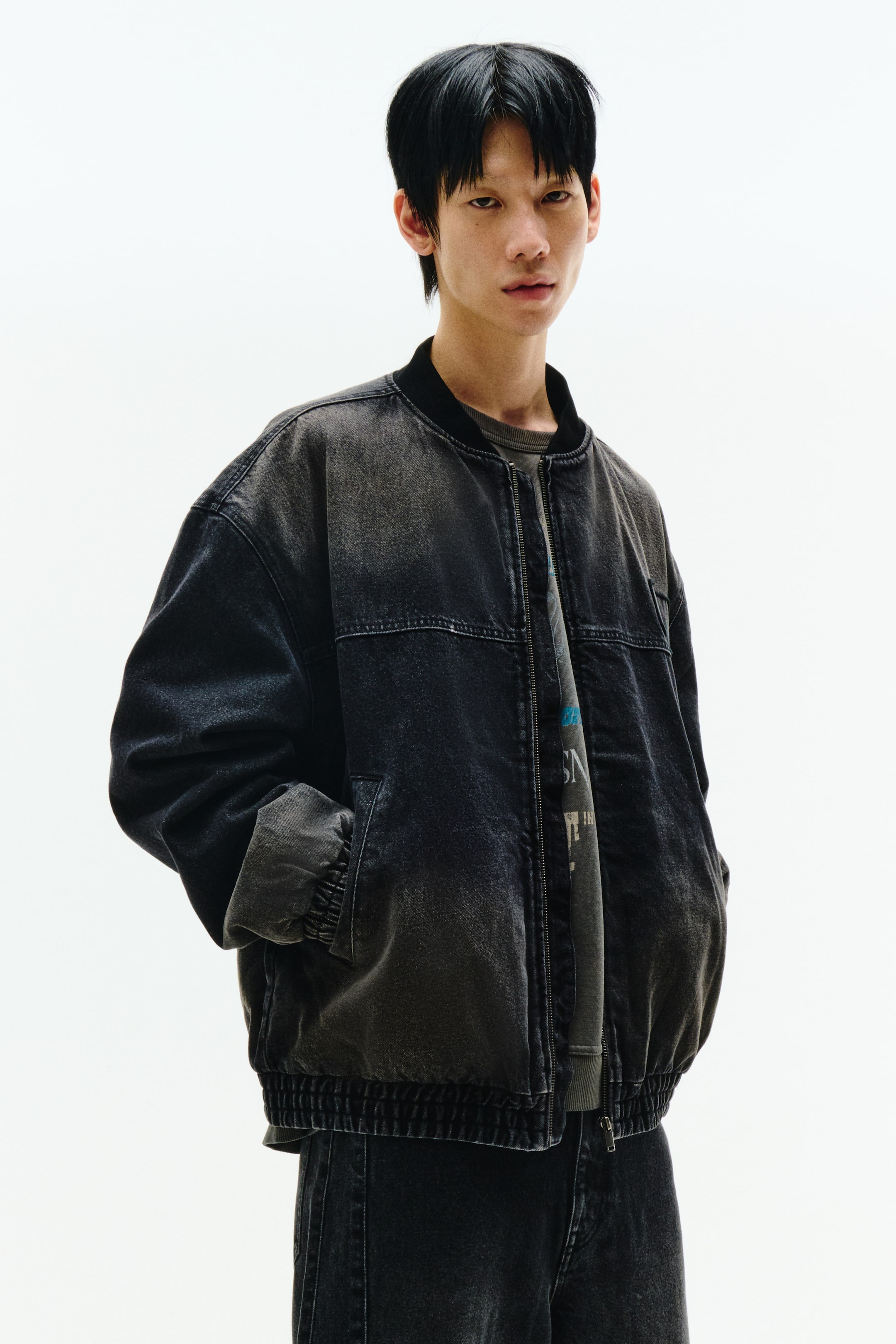 拡大画像を表示: 洗いざらしのブラックデニムボンバージャケットは、正面やや斜めからのビューで紹介されており、リブ付きカラー、フルジップクロージャー、リブ付きカフス、リブ付きヘムが特徴です。オーバーサイズのジャケットは、ダークグレーのTシャツの上に重ねられており、その裾はダークウォッシュジーンズのすぐ上に位置しています。