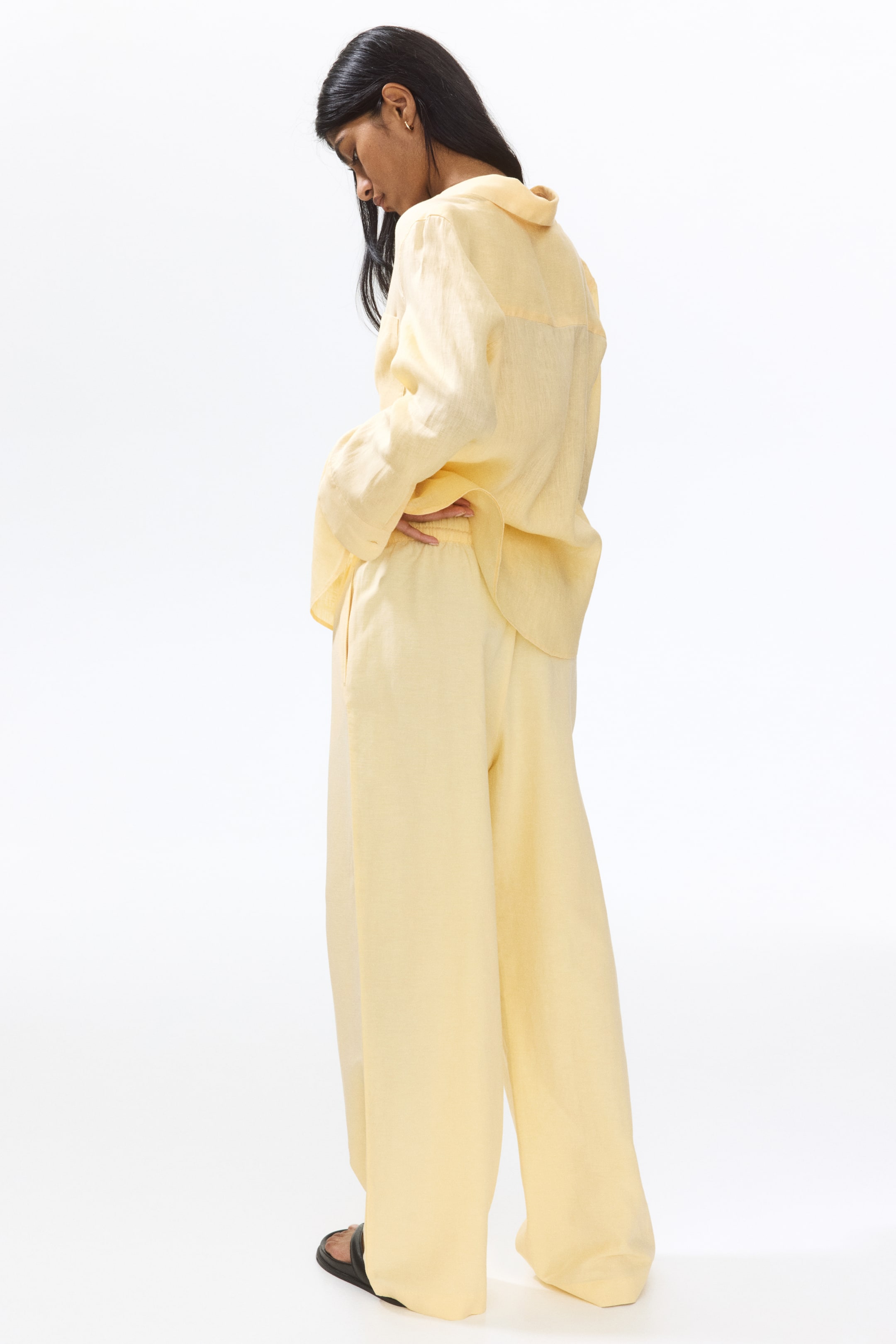 View larger image: A pale yellow long-sleeved shirt and matching wide-leg trousers are showcased from the back-left. The loose-fitting shirt features a collar and long sleeves, while the wide-leg trousers boast an elasticated waistband and side pockets.