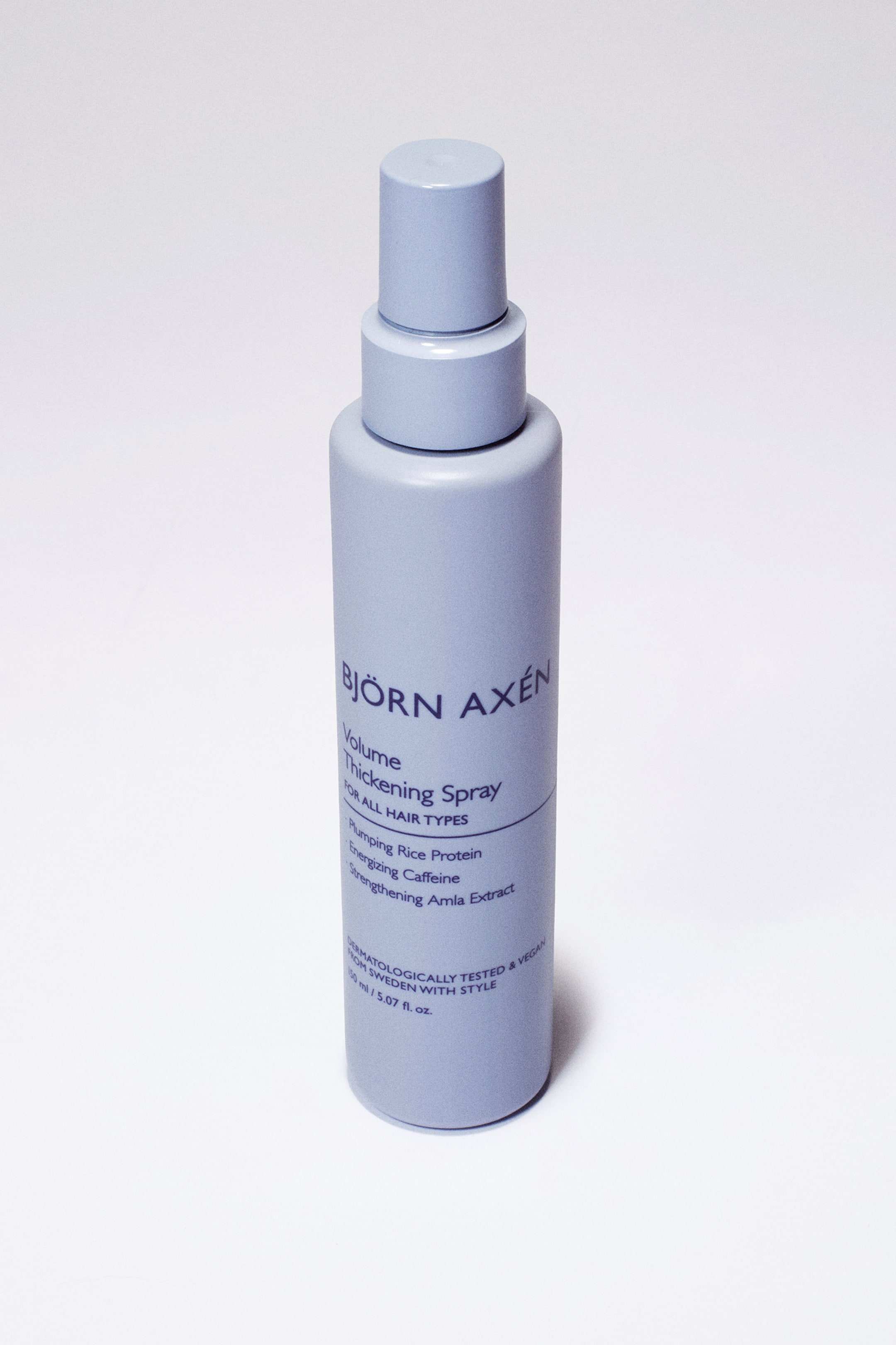 Visa större bild: En periwinklefärgad cylindrisk sprayflaska står upprätt, något lutad åt höger, och visar sin framsidesetikett. Etiketten visar varumärket "BJÖRN AXÉN" och "Volymgivande Förtjockningsspray för Alla Hårtyper", tillsammans med ingrediens- och produktinformation.