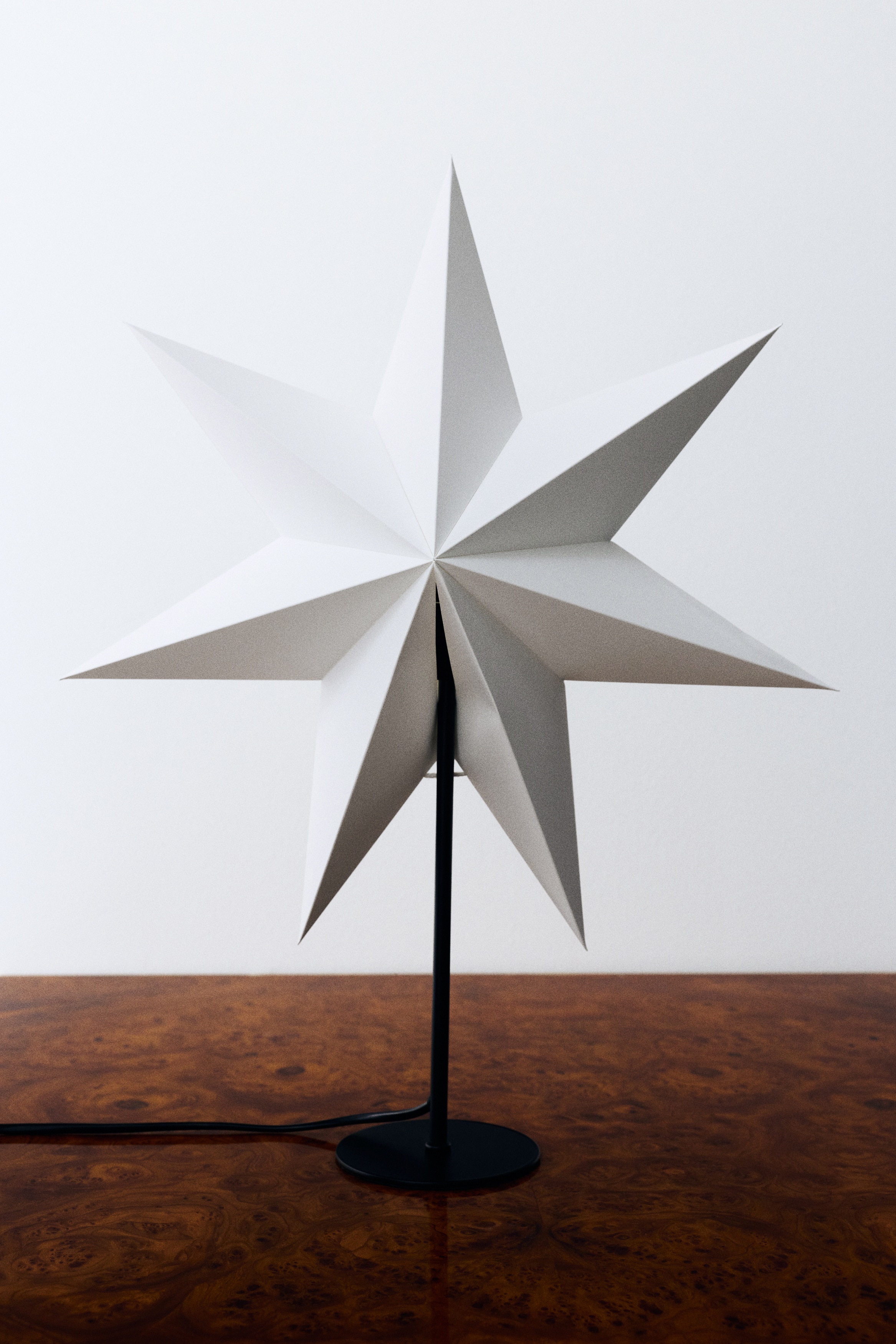 En bordslampa visar en vit, sjuddig pappersstjÃ¤rnskÃ¤rm med skarpa, tredimensionella veck, vÃ¤nd framÃ¥t pÃ¥ ett smalt svart metallstativ. Stativet reser sig frÃ¥n en platt, cirkelformad svart fot, med en svart nÃ¤tsladd som lÃ¶per ut Ã¥t sidan.