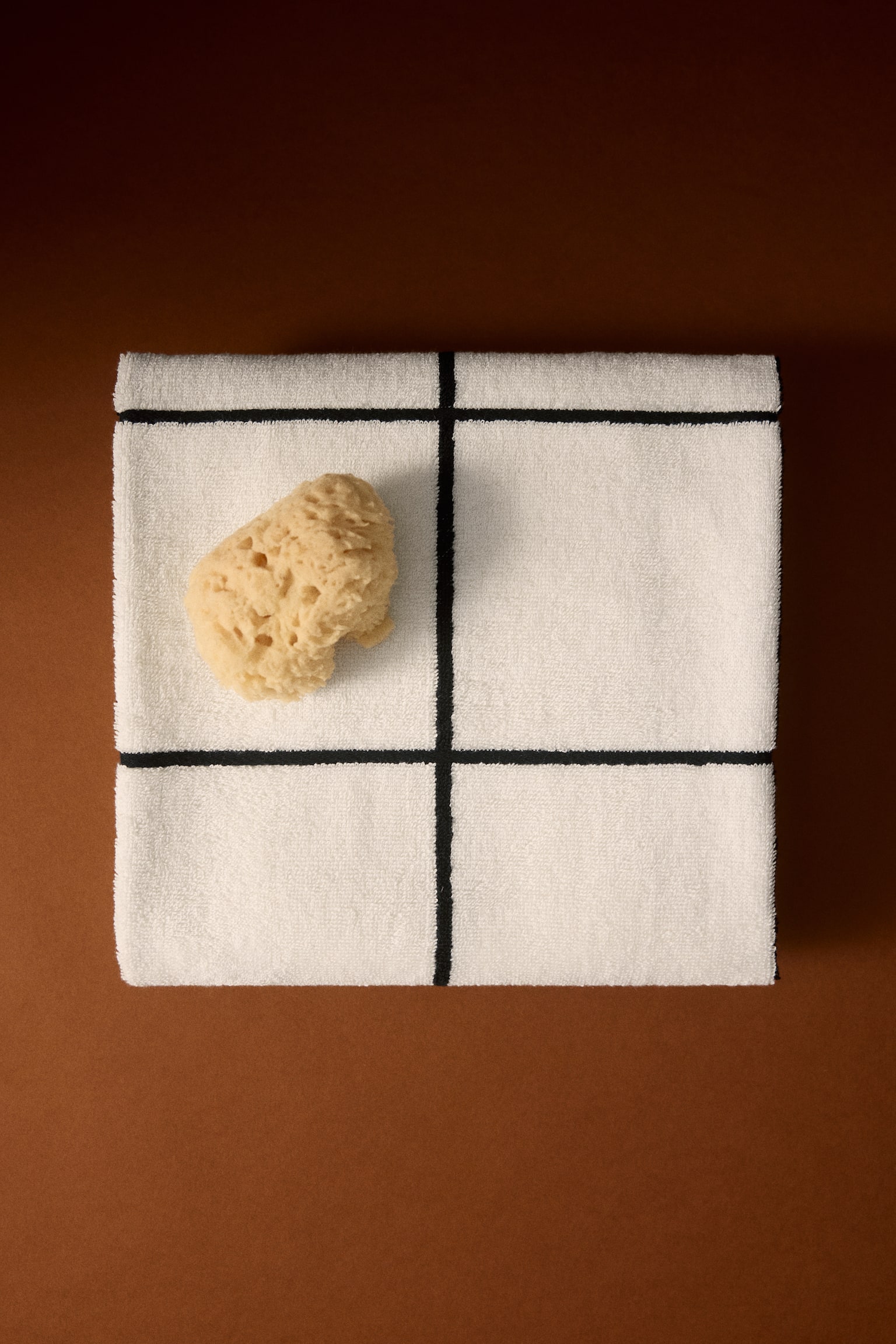 A folded cotton terry bath towel in off-white is displaying a bold black grid pattern, shown from a top-down view. A natural beige sea sponge is resting on the upper-left section of the folded towel. - 1