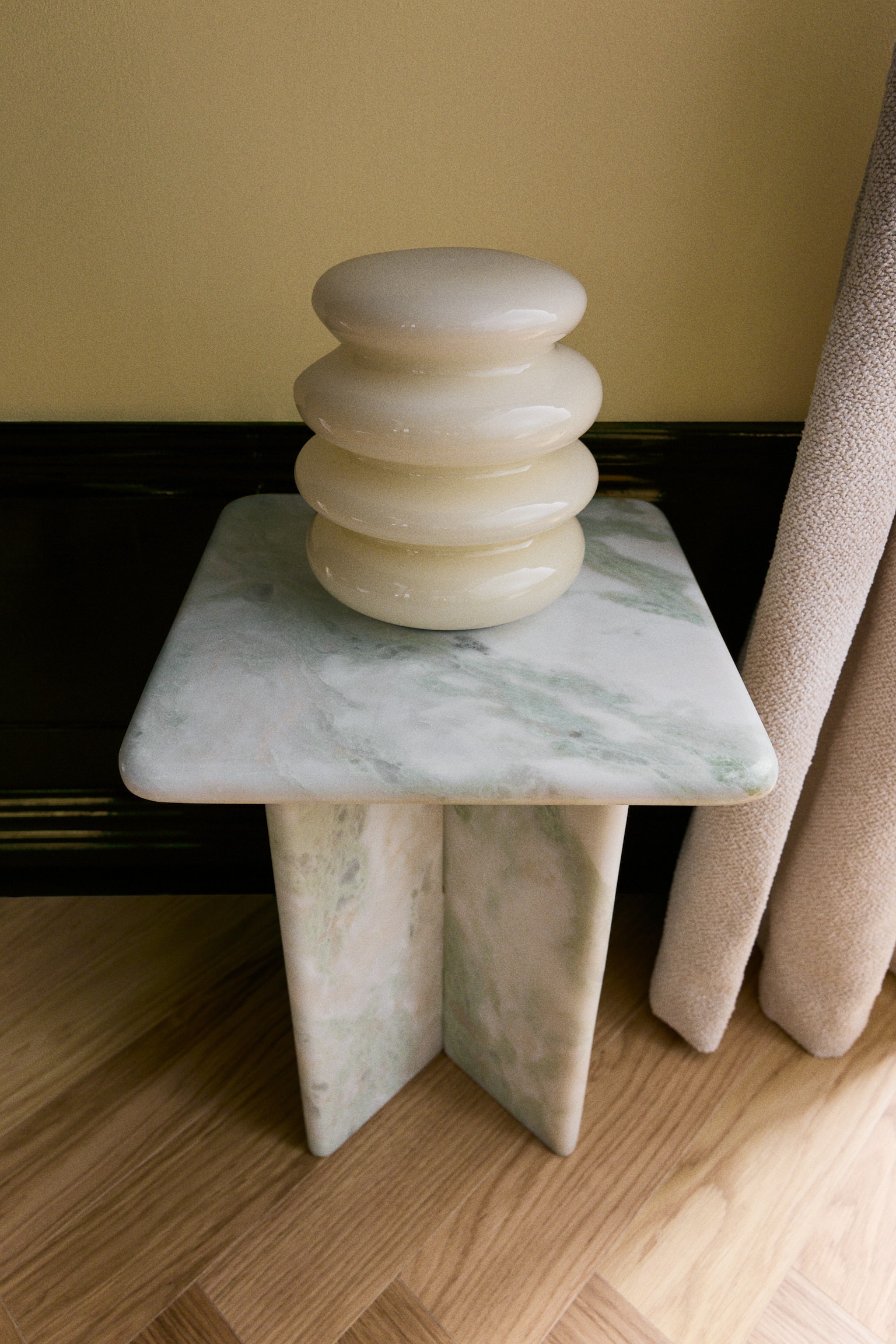 Ett kvadratiskt sidobord i marmor med ljusgröna och vita ådringar bär upp en glansig, krämfärgad våningsvas på sin plana ovansida med rundade hörn. Bordet står på en bas av korsande marmorskivor som framhäver stenens naturliga variationer. - 1