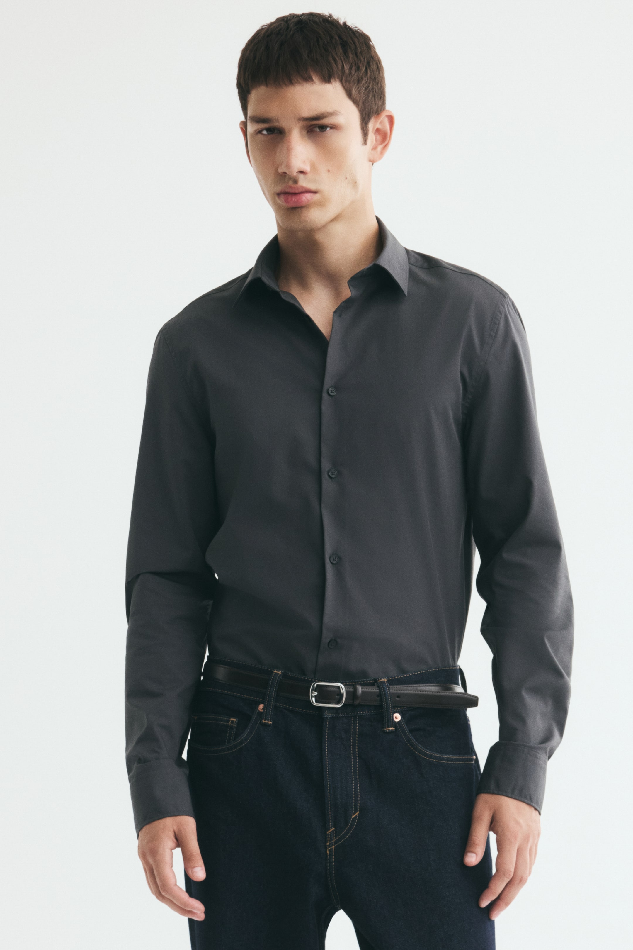 Voir une image plus grande: Le modèle porte une chemise boutonnée à manches longues gris foncé, rentrée dans un jean bleu foncé avec une ceinture noire. La chemise ajustée présente un col rabattu, une patte de boutonnage classique avec le premier bouton ouvert et des poignets boutonnés.