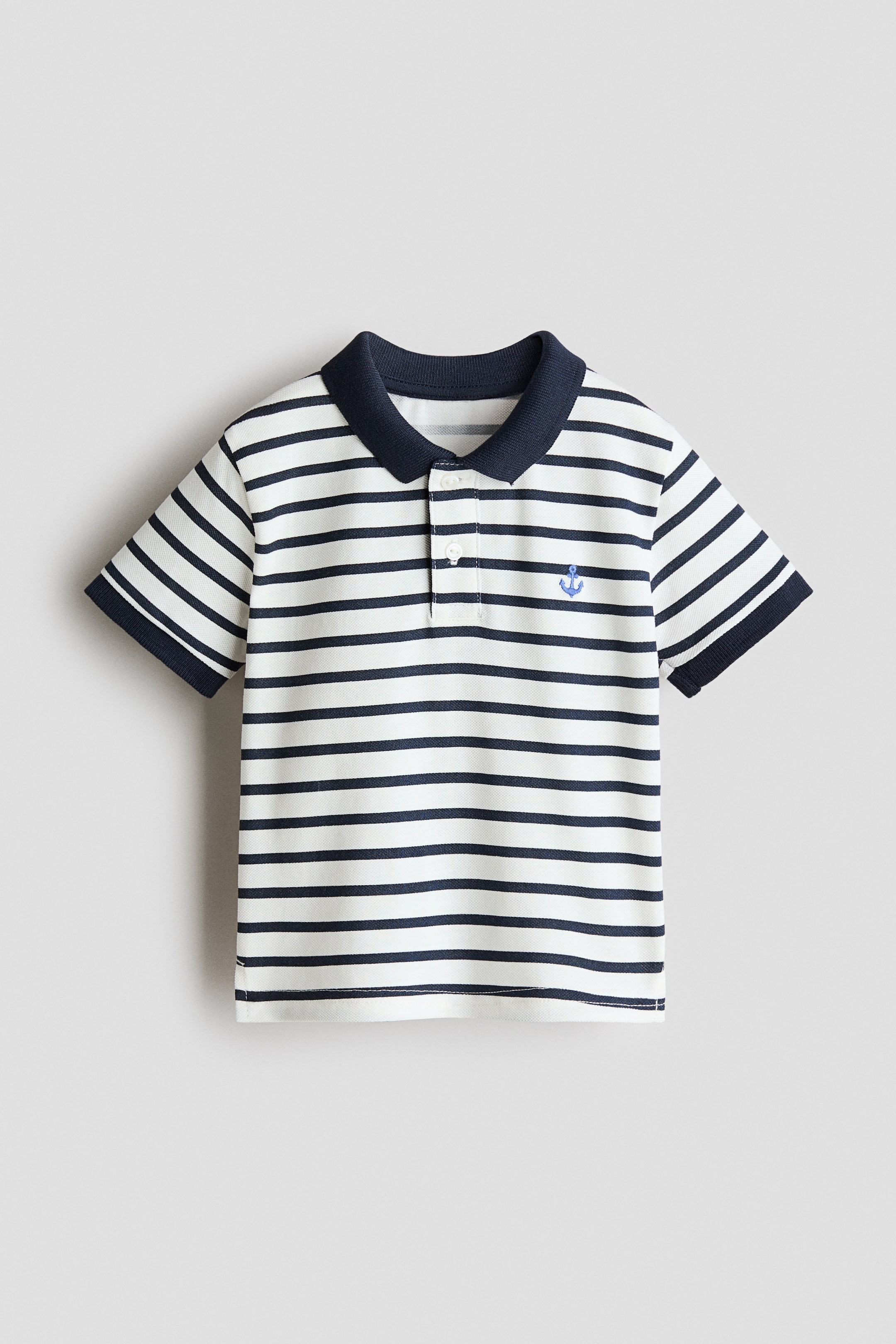Zobrazit větší obrázek: Bílé polo tričko má námořnické vodorovné pruhy, námořnický límec a dvouknoflíkové zapínání s bílými knoflíky. Malá modrá kotva je vyšitá na levé straně hrudi a krátké rukávy jsou zakončeny námořnickými žebrovanými lemy.
