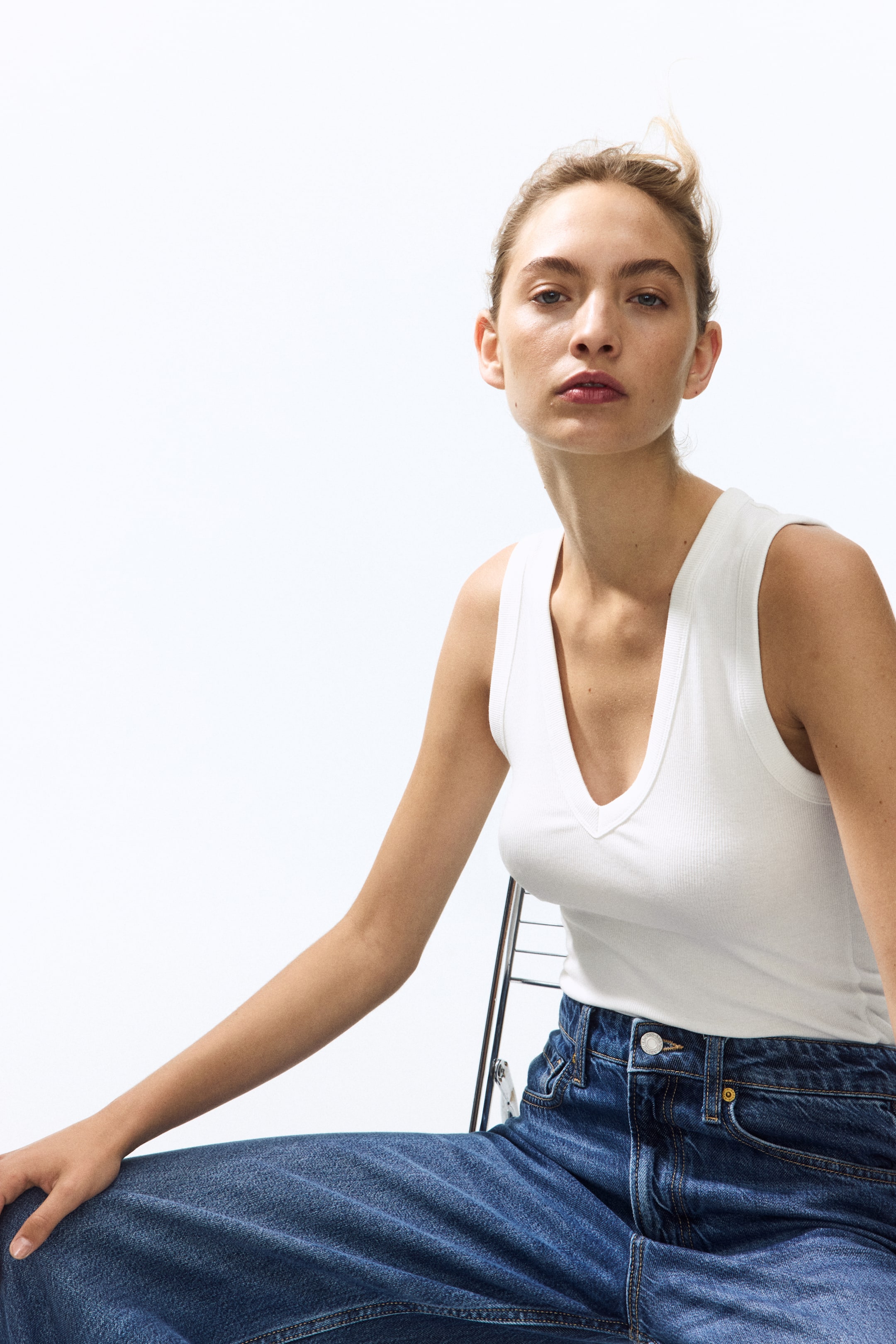 View larger image: A white ribbed tank top with a deep V-neck is worn, paired with dark blue, wide-leg denim jeans featuring a visible button closure and contrast stitching.