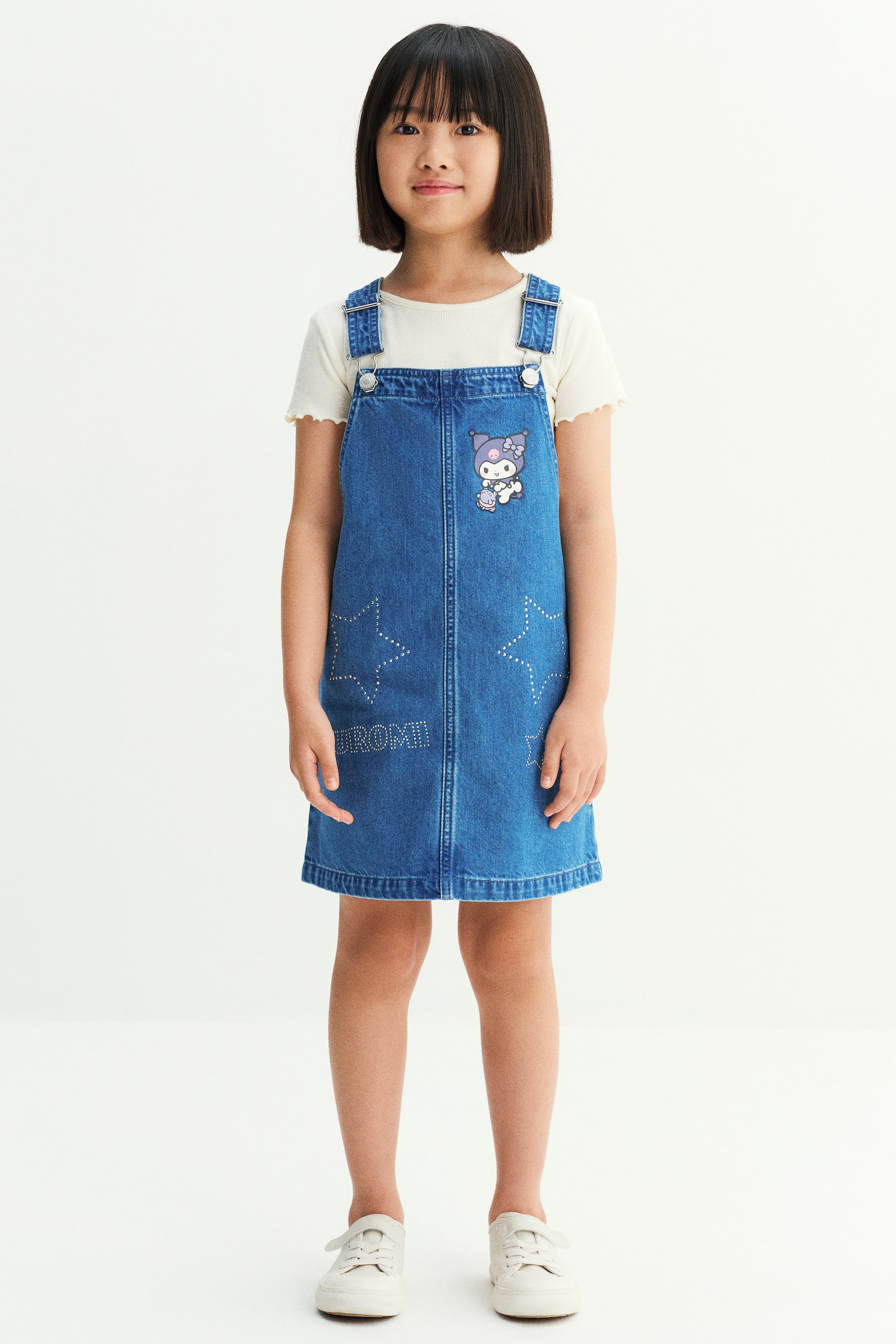 Xem ảnh lớn hơn: Người mẫu đang mặc một bộ váy yếm, gồm áo thun jersey cotton gân màu kem được phối bên trong một chiếc váy yếm denim màu xanh. Áo thun có cổ tròn và bo tay may vắt sổ, trong khi chiếc váy có dây vai điều chỉnh được với khóa kim loại, họa tiết nhân vật hoạt hình ở ngực và thiết kế ngôi sao đính đá rhinestone ở mặt trước.