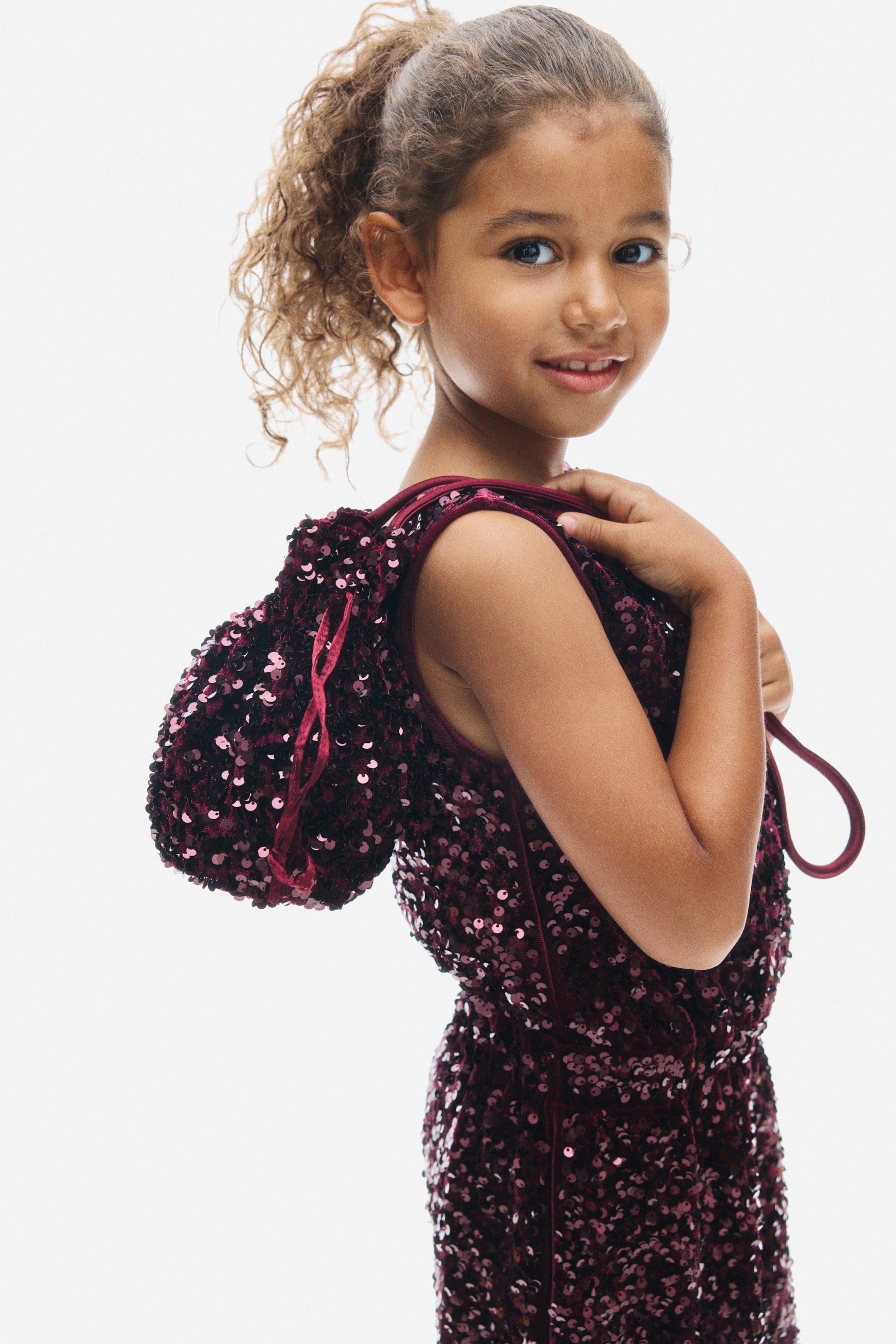 View larger image: A sleeveless burgundy playsuit in sequin-covered velour is shown from a side profile facing right, showcasing its shimmering texture and velour-trimmed armhole. A matching sequined drawstring bag is hanging over the shoulder, coordinating with the playsuit.
