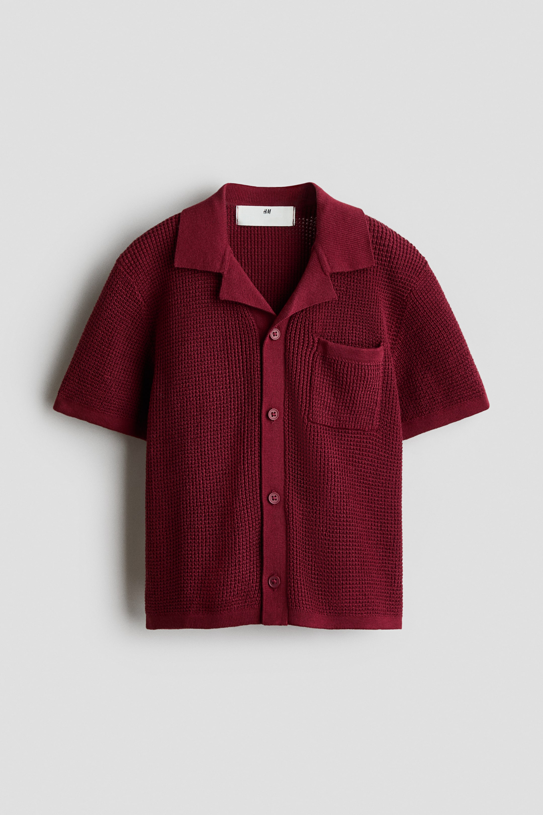 View larger image: A dark red waffle-knit polo shirt is laid flat, facing front. It features a spread collar, short sleeves, a full button placket with matching buttons, and a single patch pocket on the left chest.