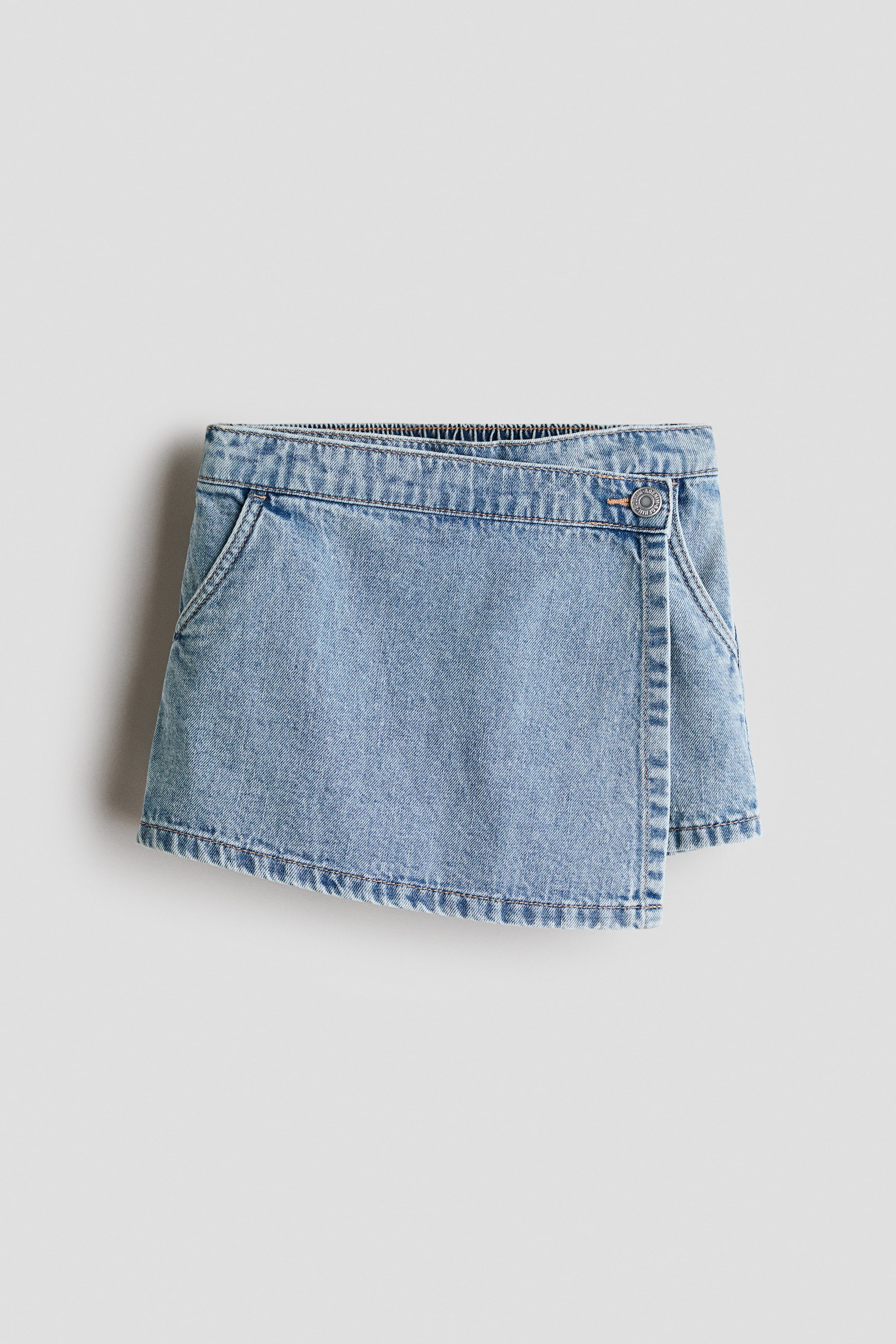 View larger image: A light blue denim skort is hanging, showcasing a wrap-style front with an asymmetrical hem. It features a single metal button closure on the right, two slanted front pockets, and an elasticated back waistband.