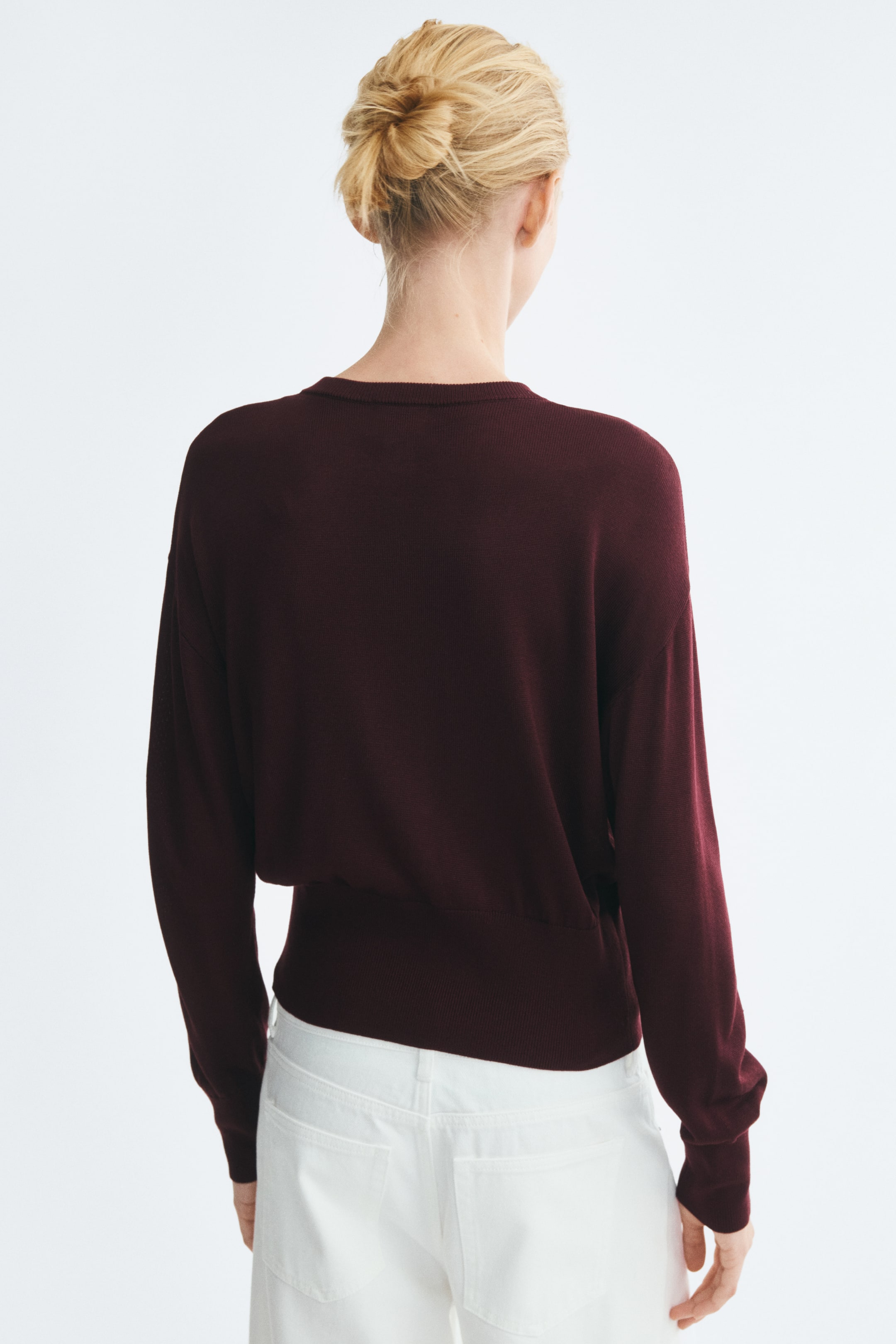 View larger image: A burgundy knit cardigan is worn by the model, shown from the back. The cardigan features a round neckline, dropped shoulders, long sleeves with ribbed cuffs, and a wide ribbed waistband, and is paired with white trousers.