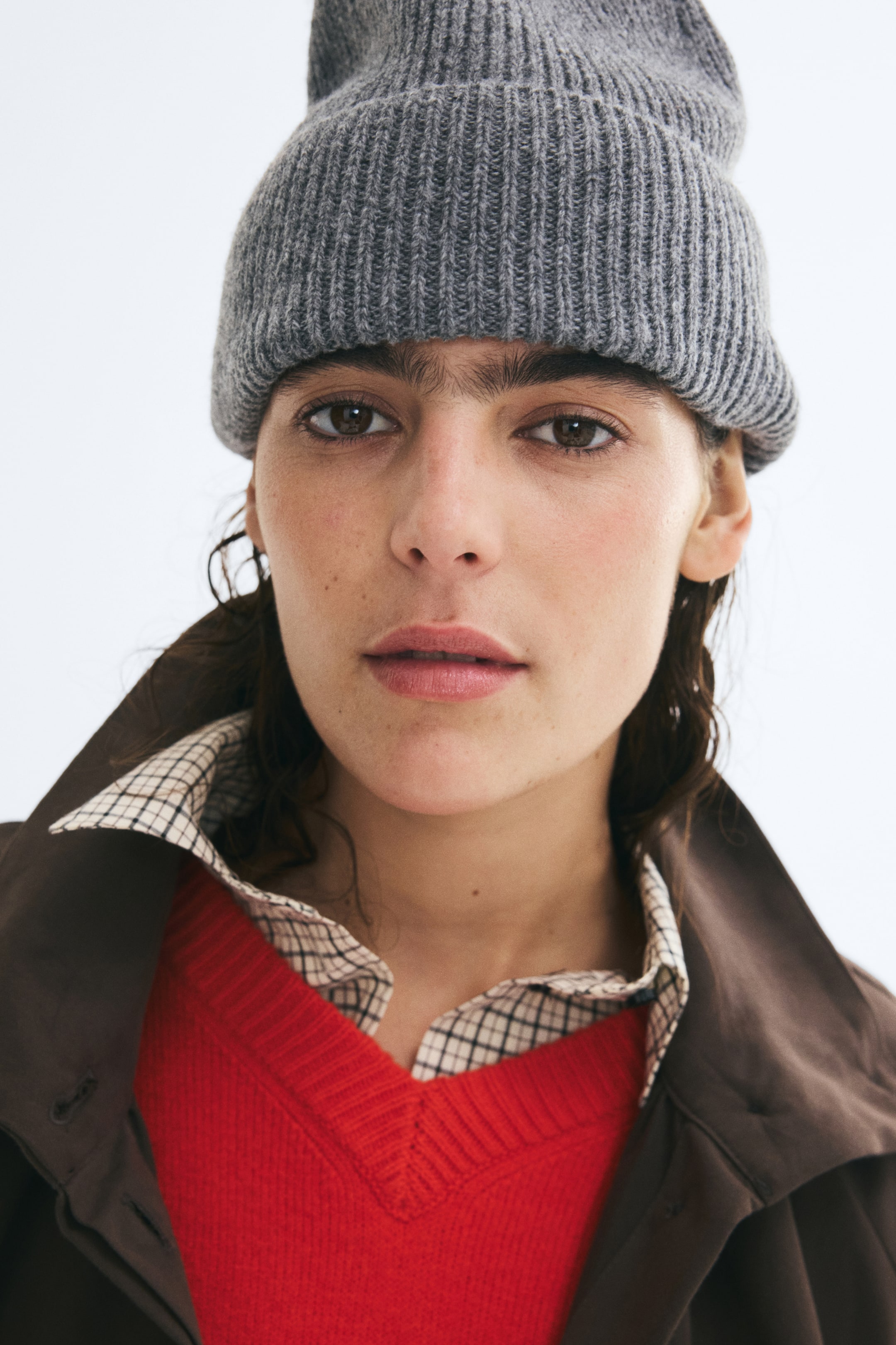Ver imagen más grande: Una gorra gris de punto acanalado con vuelta en el borde la lleva puesta la modelo. La gorra se combina con un suéter rojo de cuello en V, superpuesto sobre una camisa de cuello a cuadros y una chaqueta marrón.