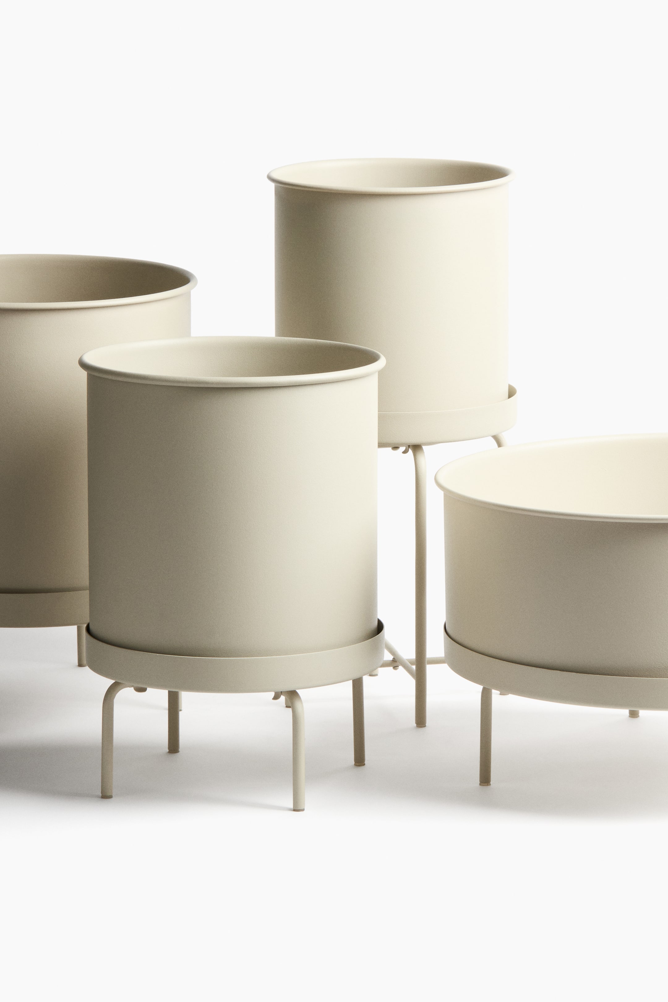 Ampliar la imagen: Cuatro macetas cilíndricas de metal están colocadas sobre soportes metálicos a juego, todas en un beige claro uniforme. Cada soporte cuenta con un platillo que sostiene la maceta, y se presentan en distintos tamaños y alturas, con un soporte notablemente más alto.