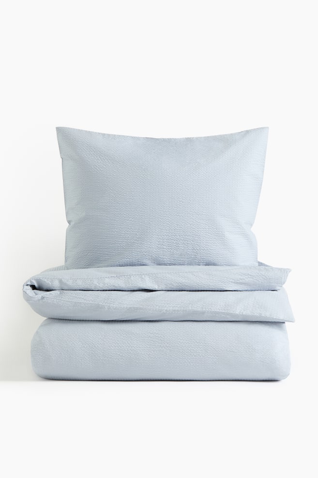 A light blue seersucker duvet cover set is displayed from the front, showcasing a pillowcase standing upright behind a neatly folded duvet cover. Both items exhibit the characteristic puckered texture of seersucker fabric. - 1