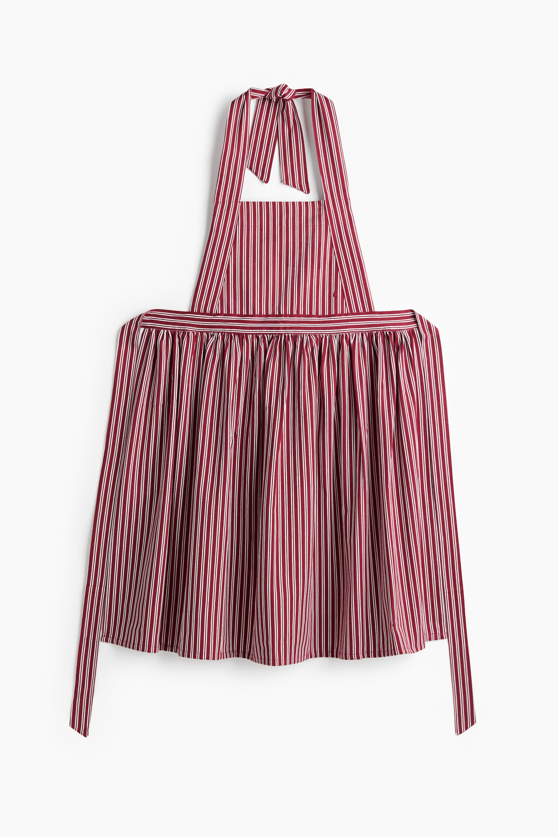 View larger image: A red and white vertically striped bib apron is hanging flat, facing front. It features a halter neck strap tied in a bow at the top, a gathered waistline that creates a draped skirt, and long matching waist ties hanging from the sides.