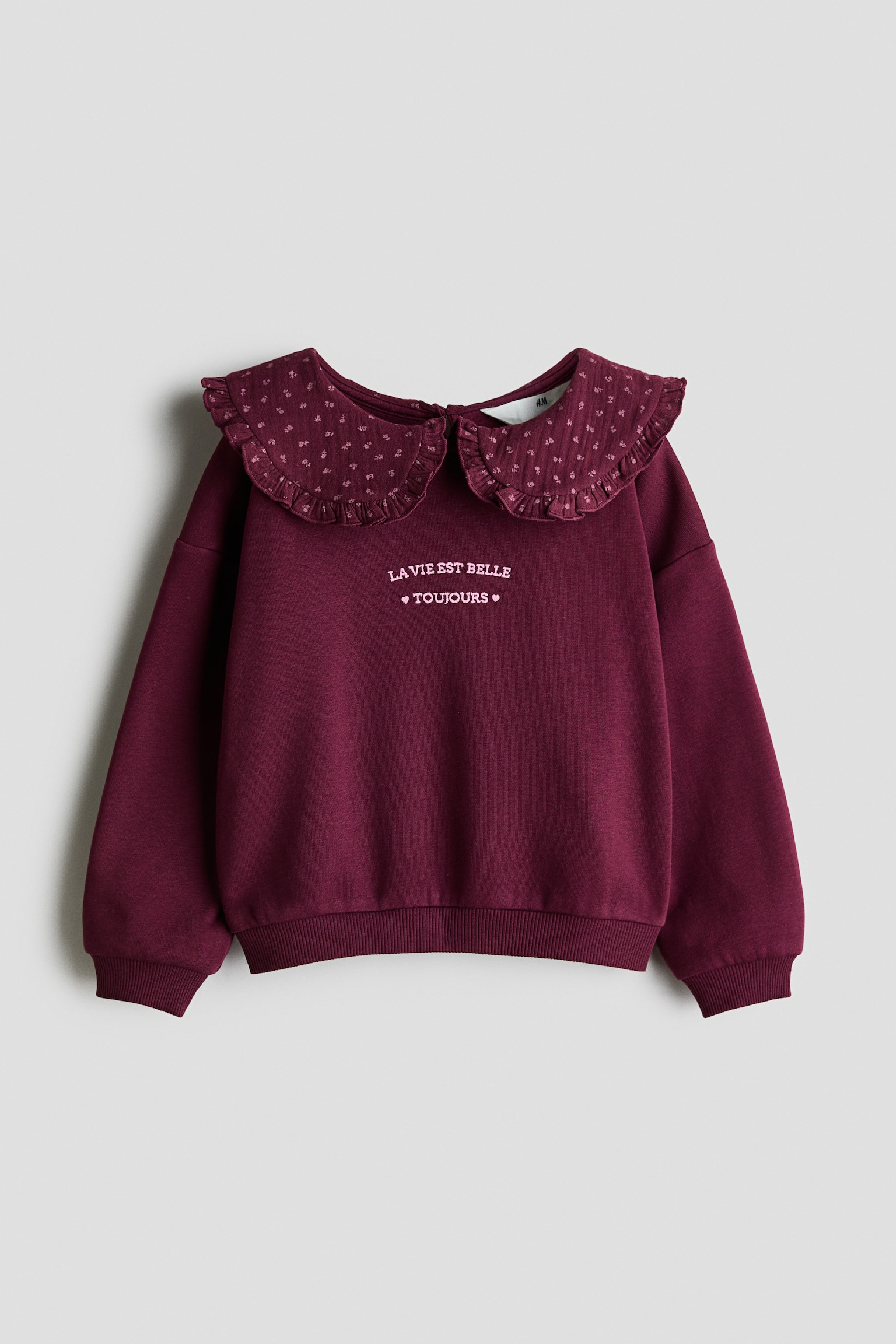 A front-facing burgundy sweater in brushed fleece fabric features a ruffled, patterned Peter Pan collar and white embroidered text on the chest. The design features dropped shoulders, long sleeves with ribbed cuffs, and a ribbed hem. - 1